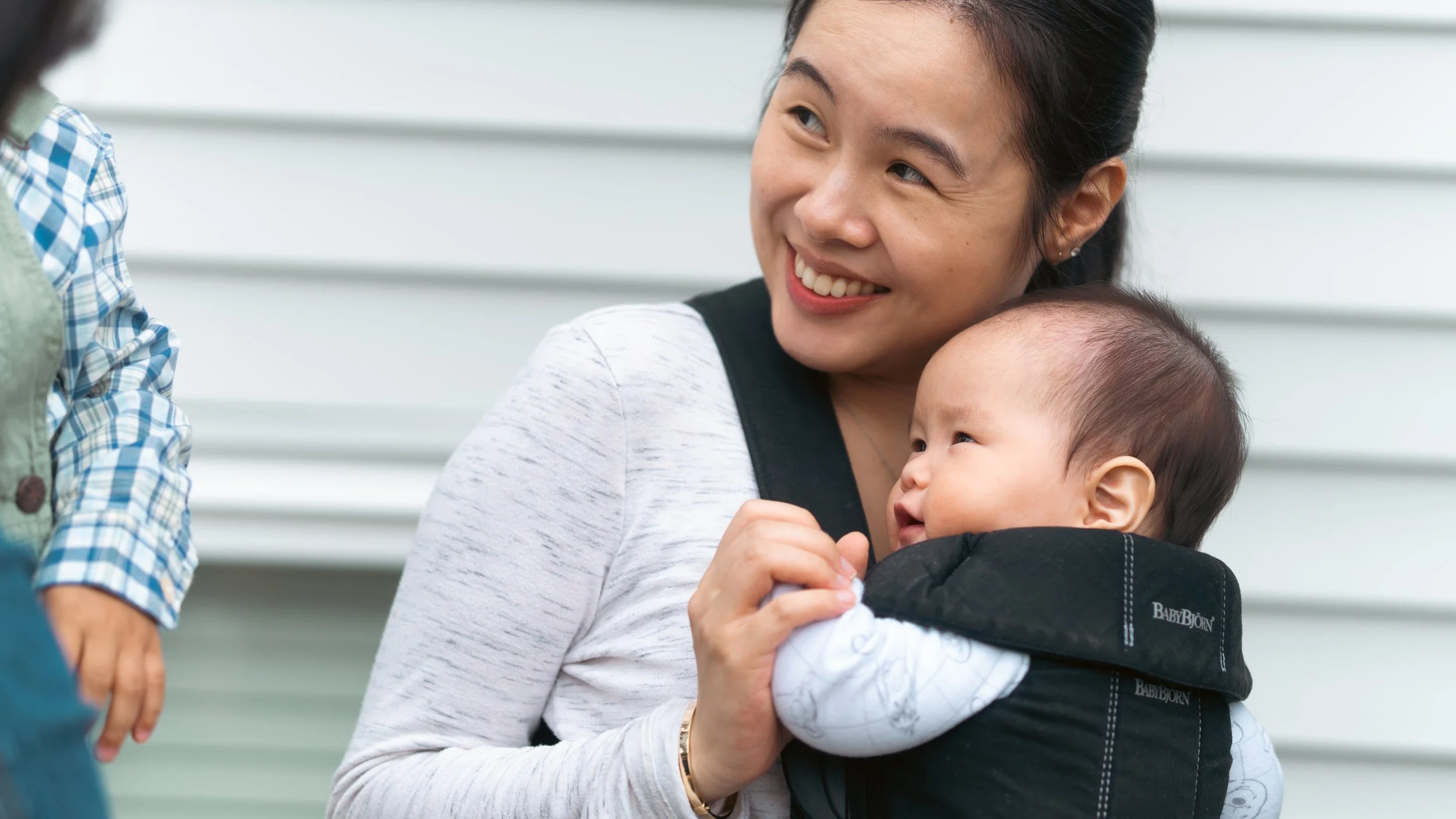 A smiling woman holding a baby in a black carrier in front of a white wall.