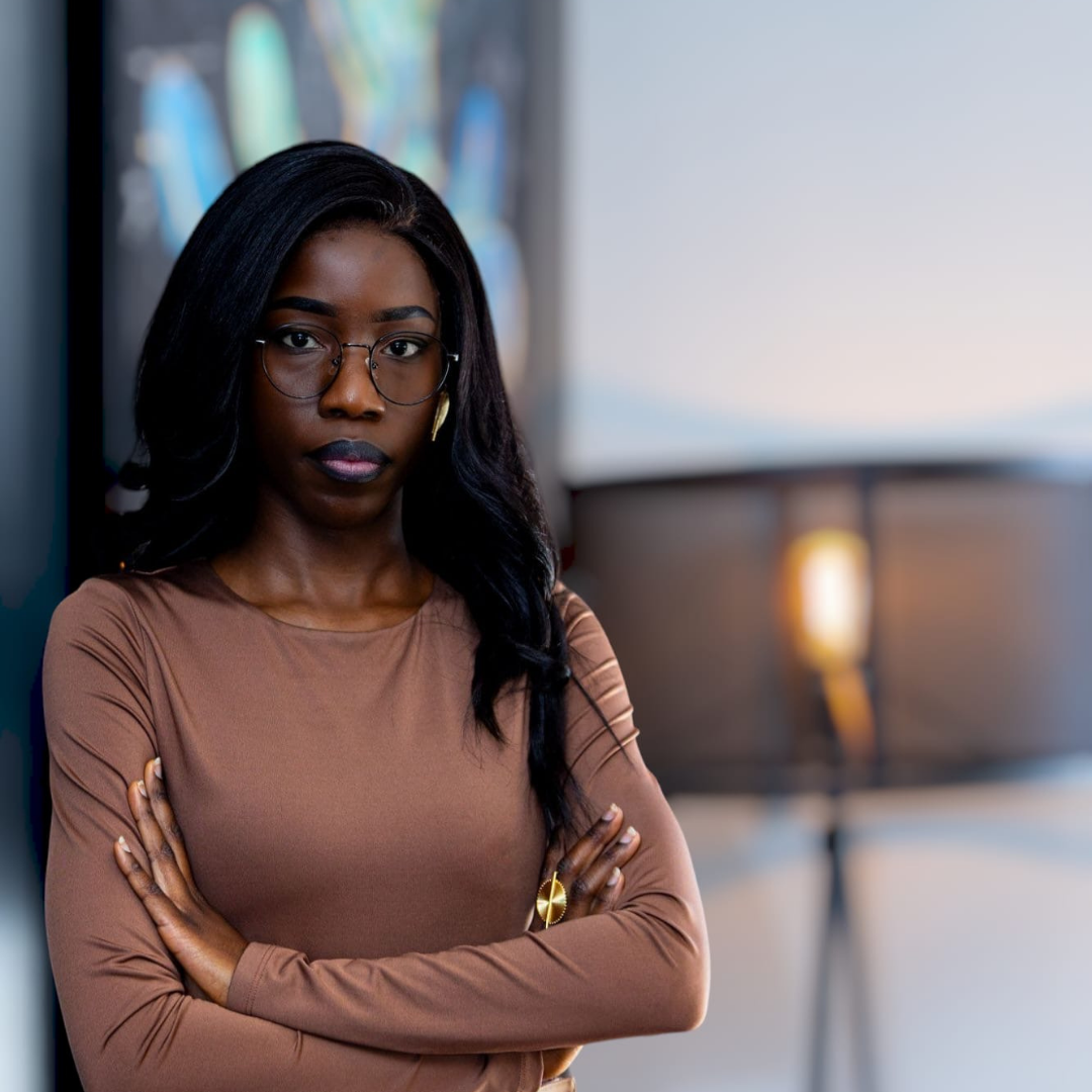 Femme noire portant des lunettes, en tenue beige, debout avec les bras croisés dans un environnement moderne, probablement un bureau.