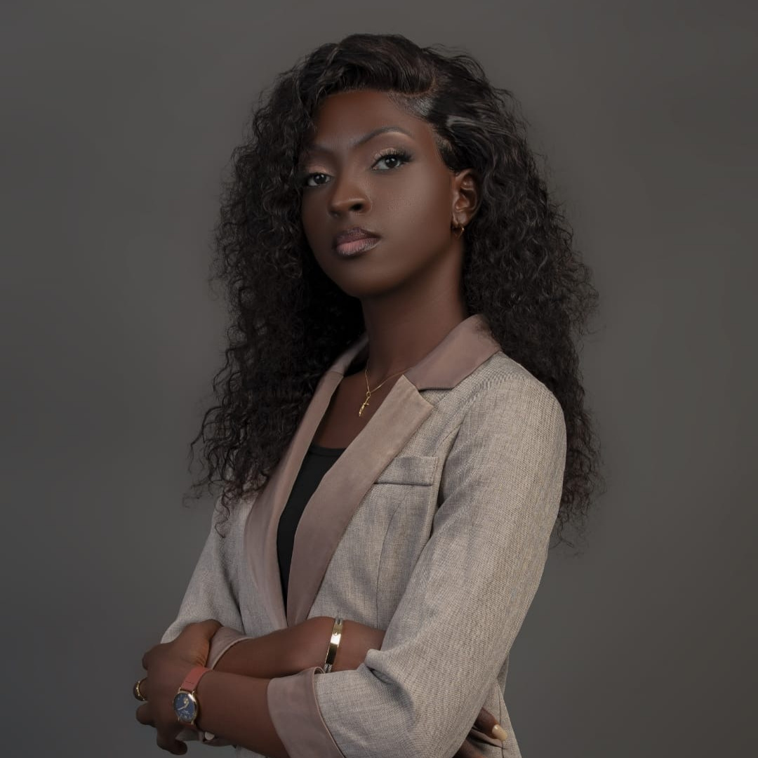 Portrait d'une femme noire avec des cheveux bouclés foncés, portant une veste beige et un collier avec une croix, posant avec les bras croisés sur un fond gris.