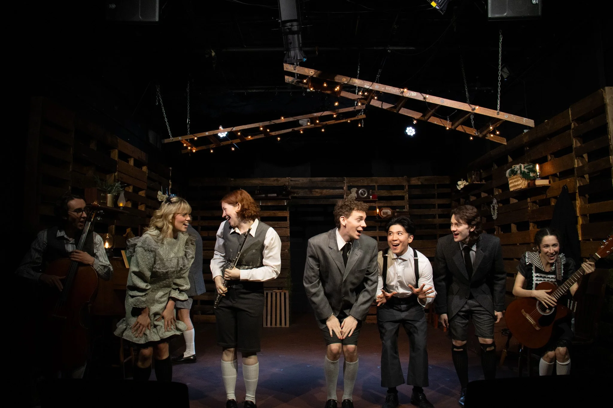 The cast of a theatrical play on stage. Seven actors are in costume, some holding musical instruments, in a rustic set with wooden pallets and warm lighting.