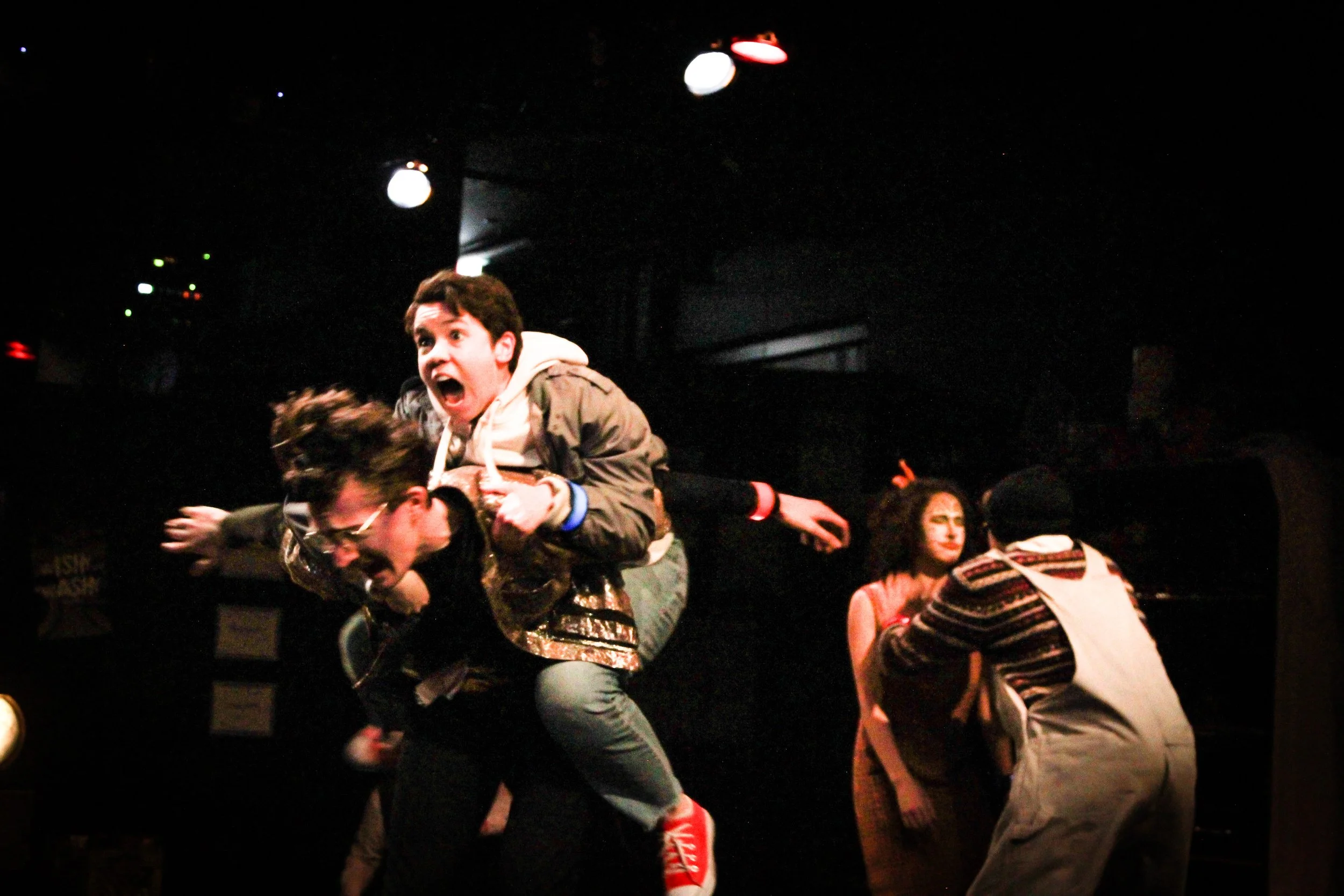Young people performing on stage with dramatic expressions; one person is giving another a piggyback ride, while others are engaged in different actions.