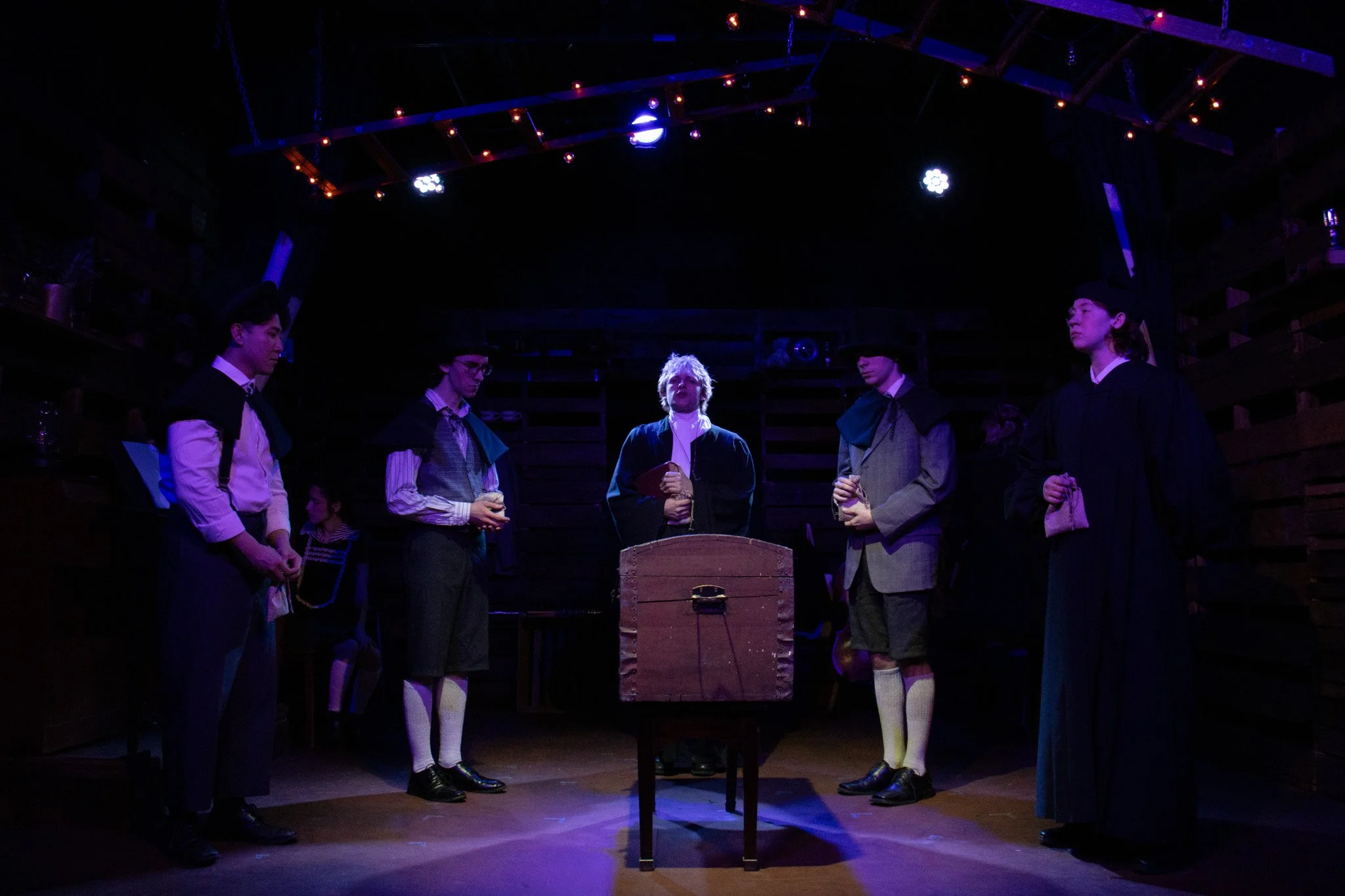 A theatrical scene with six actors dressed in period costumes standing on a stage with a wooden backdrop, illuminated by purple stage lighting. The actors are positioned around a small wooden table, and one actor in the center appears to be speaking