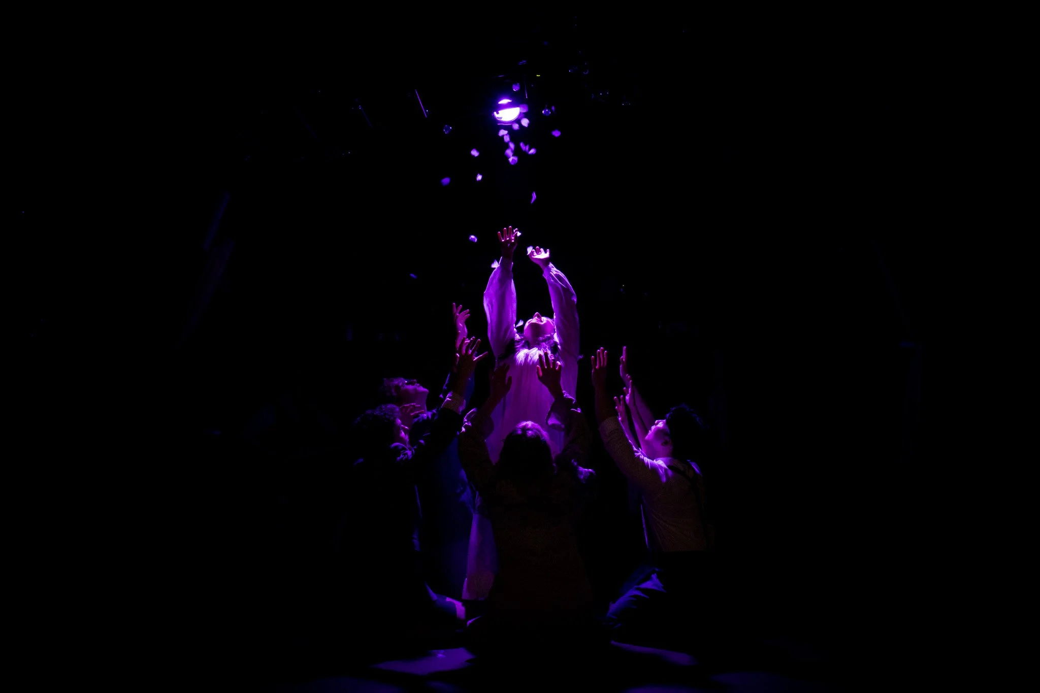 A group of people reaching towards a person in a purple-lit dark space, with the person in the center bathed in light, reaching upward as if receiving or embracing the light.