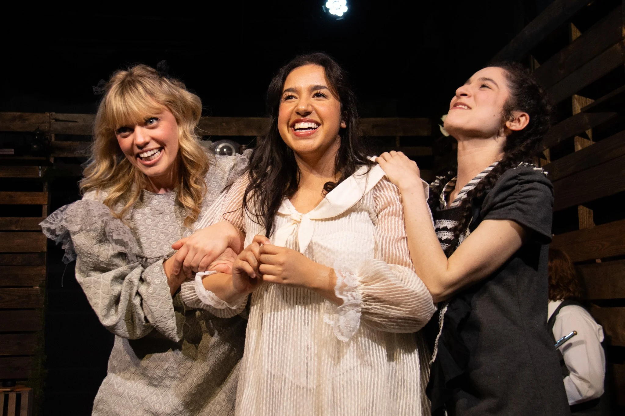 Three women standing close together, smiling and laughing, with two women on either side of a woman in the middle. They are inside with dark wooden walls in the background.