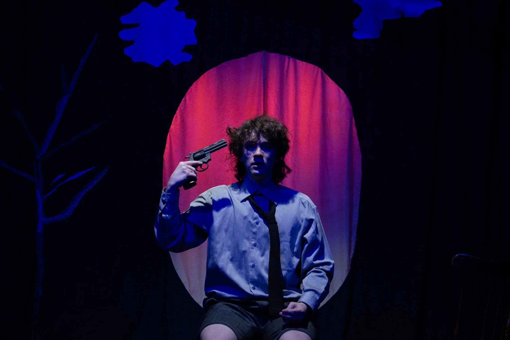 A young person with curly hair seated on stage, holding a gun to their head, with a red curtain behind them and a dark background featuring a blue tree silhouette and clouds.