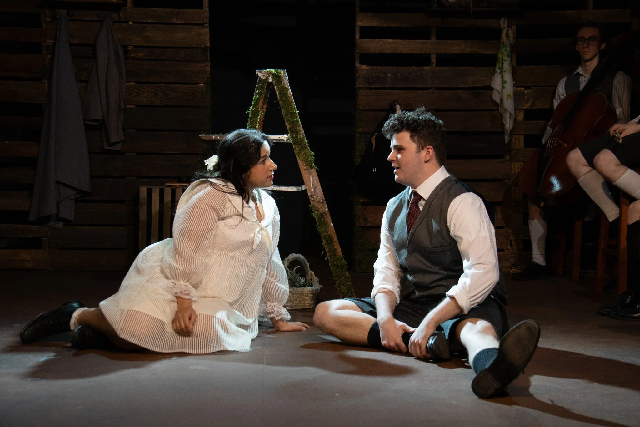 Two actors kneel on the stage, facing each other, during a theatrical performance. One woman wears a white nightgown, and one man wears a vest with rolled-up shirt sleeves and shorts. A musical ensemble with a cellist is visible in the background.