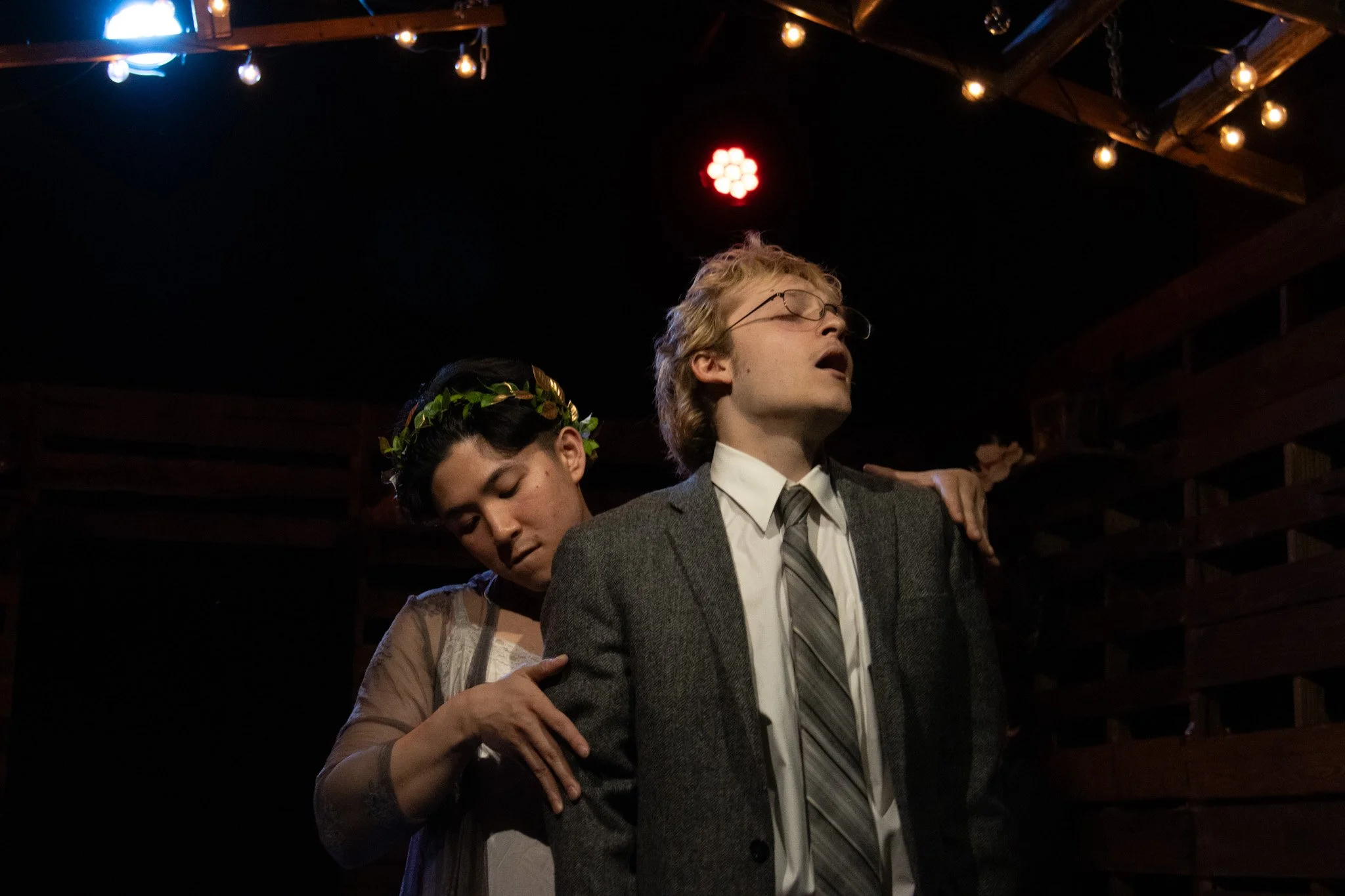 Two actors performing on stage, one man with glasses and a gray suit, the other woman with a flower crown, appear to be in a theatrical scene under dim lighting with string lights overhead.