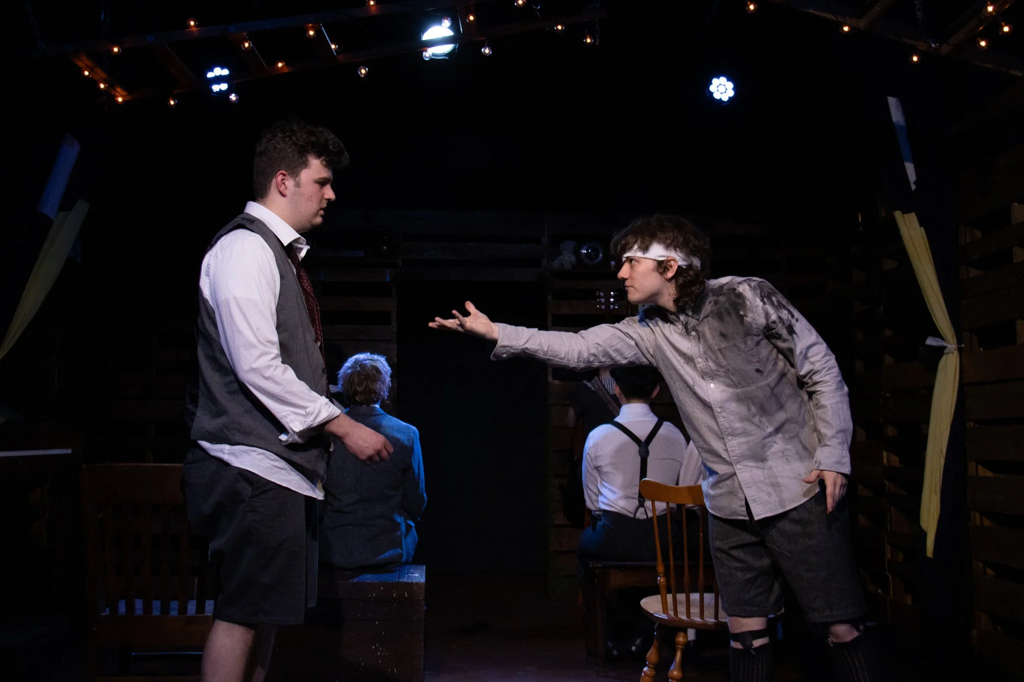 Two young men are engaged in an intense conversation on stage during a theatrical performance, with a group of people seated in the background and stage lights overhead.