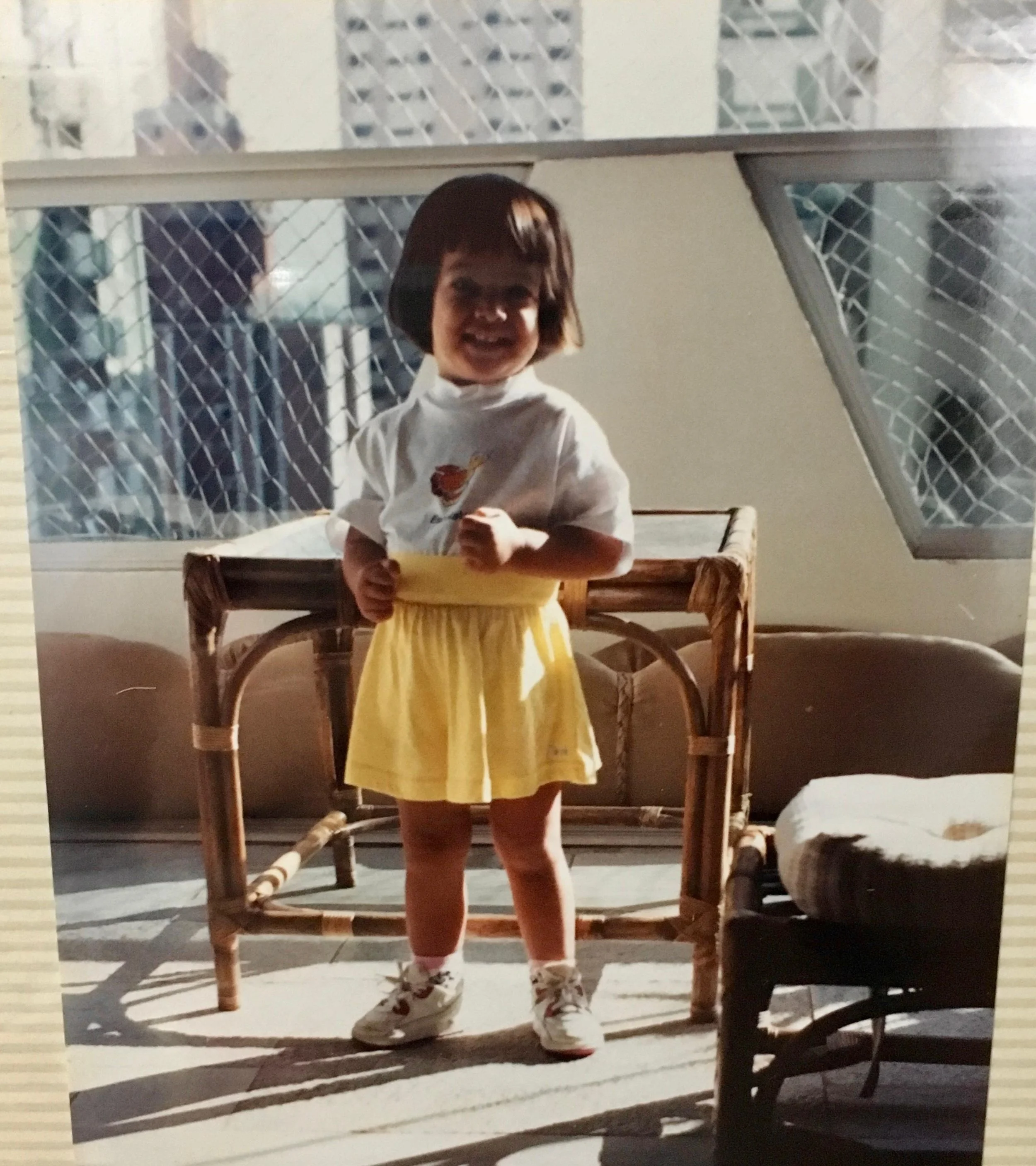 Myself at around 4 years old. Small girl with bowl haircut, dark hair, cheerful smile and chubby cheeks wearing a yellow skirt.
