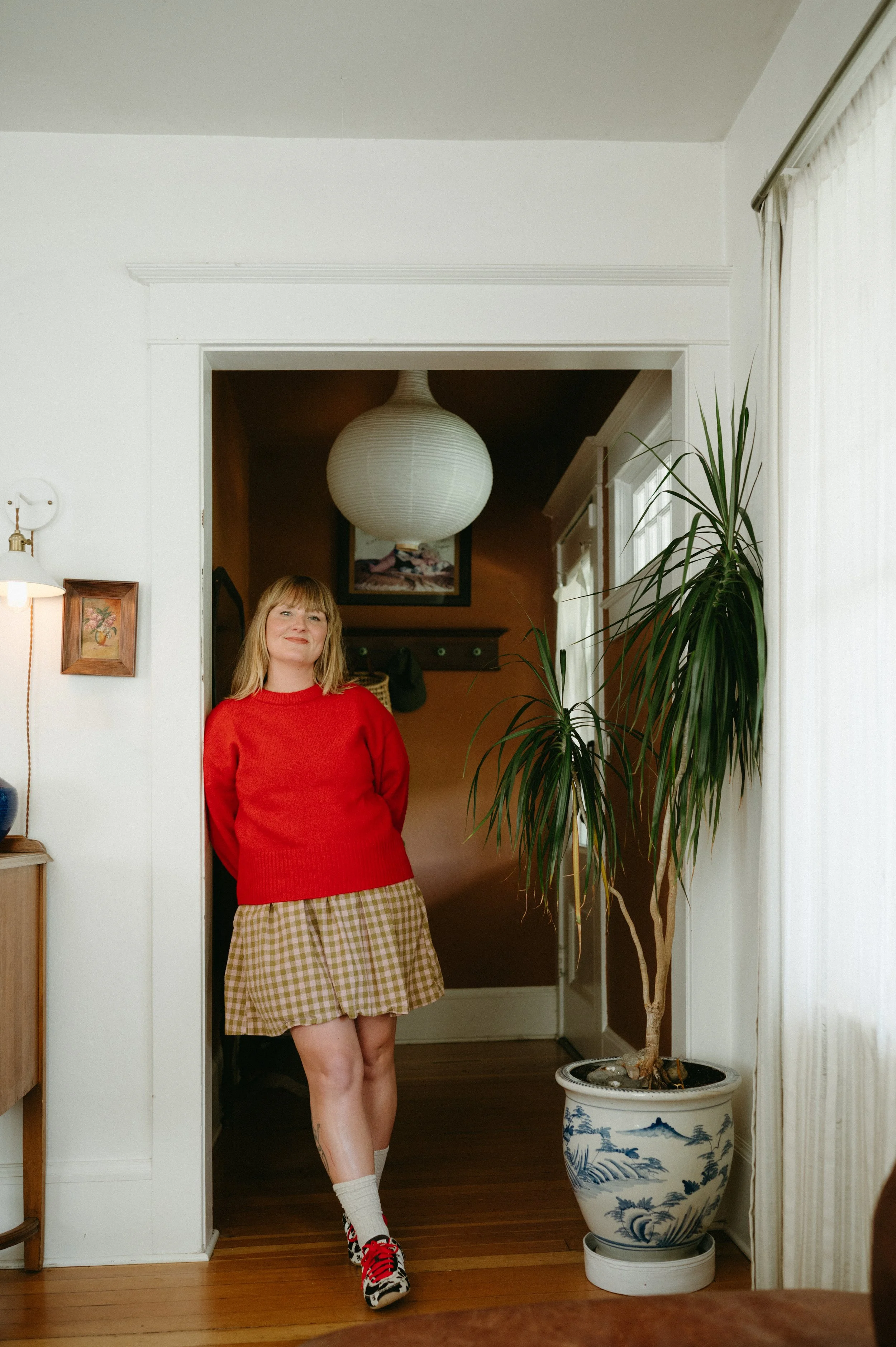A woman with blonde hair wearing a red sweater, checkered skirt, and red and black sneakers standing in a doorway next to a tall potted plant.