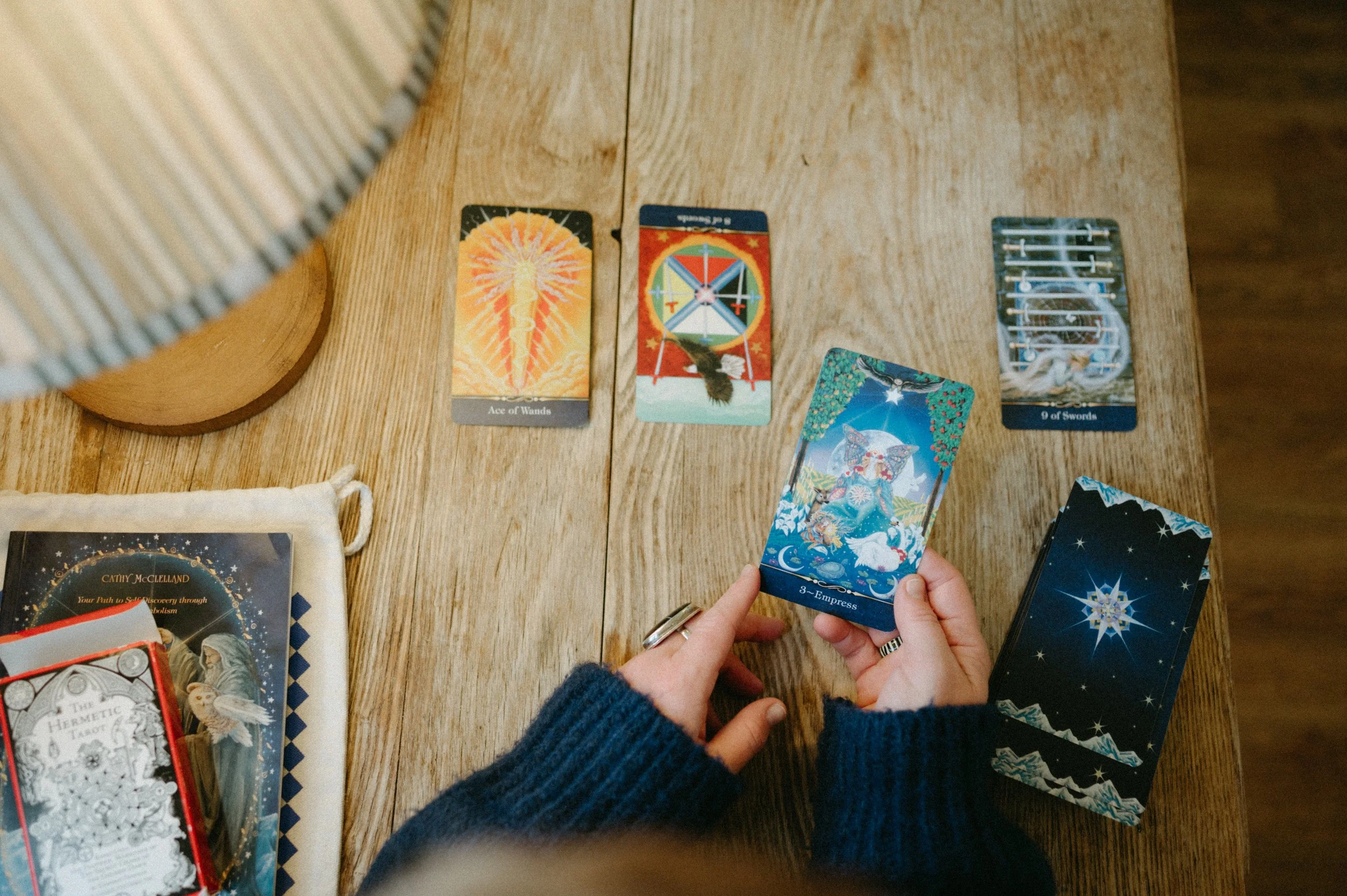 A person holding a tarot card labeled '3 - Empress' in front of a spread of tarot cards on a wooden table. Other cards and tarot books are also visible on the table.