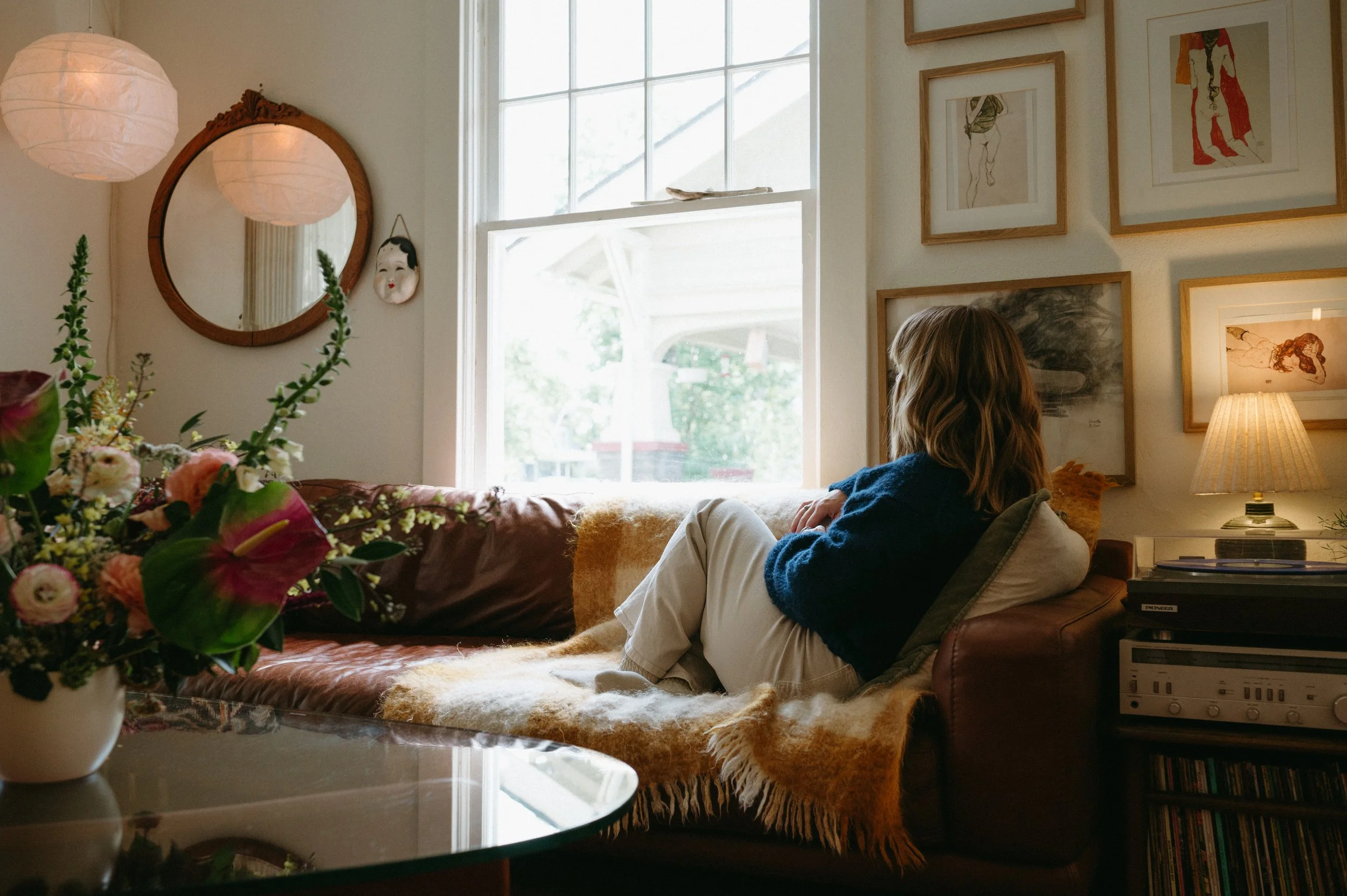 A person with long hair sitting on a brown leather couch in a living room, looking towards the window. The room is decorated with framed artwork on the wall, a lamp, and a large flowers bouquet on the table.