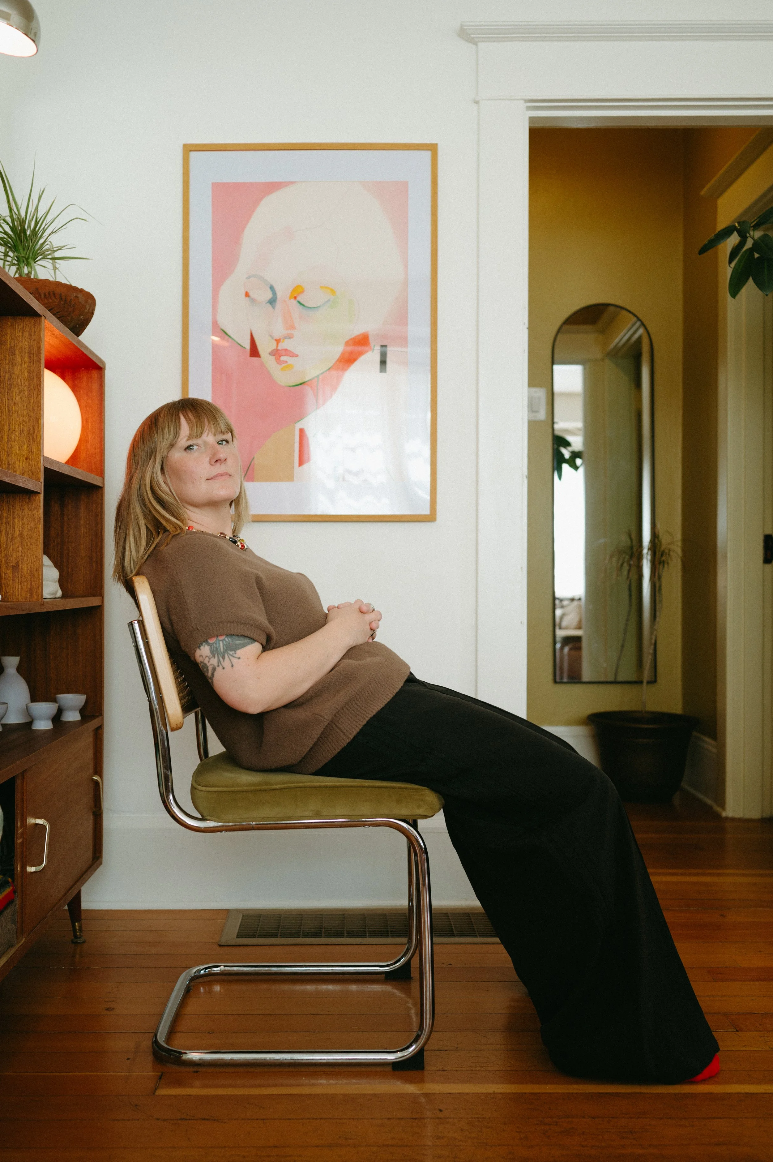 A woman with light brown hair and tattoos on her arm sitting on a chair with her back leaning against the wall, in a room with wooden flooring and white walls. She is wearing a brown top and black pants. Behind her is a colorful abstract portrait of a face on the wall, and to her left is a wooden shelf with decorative objects and a potted plant. There is a doorway to a yellow room with another mirror and a plant in the background.