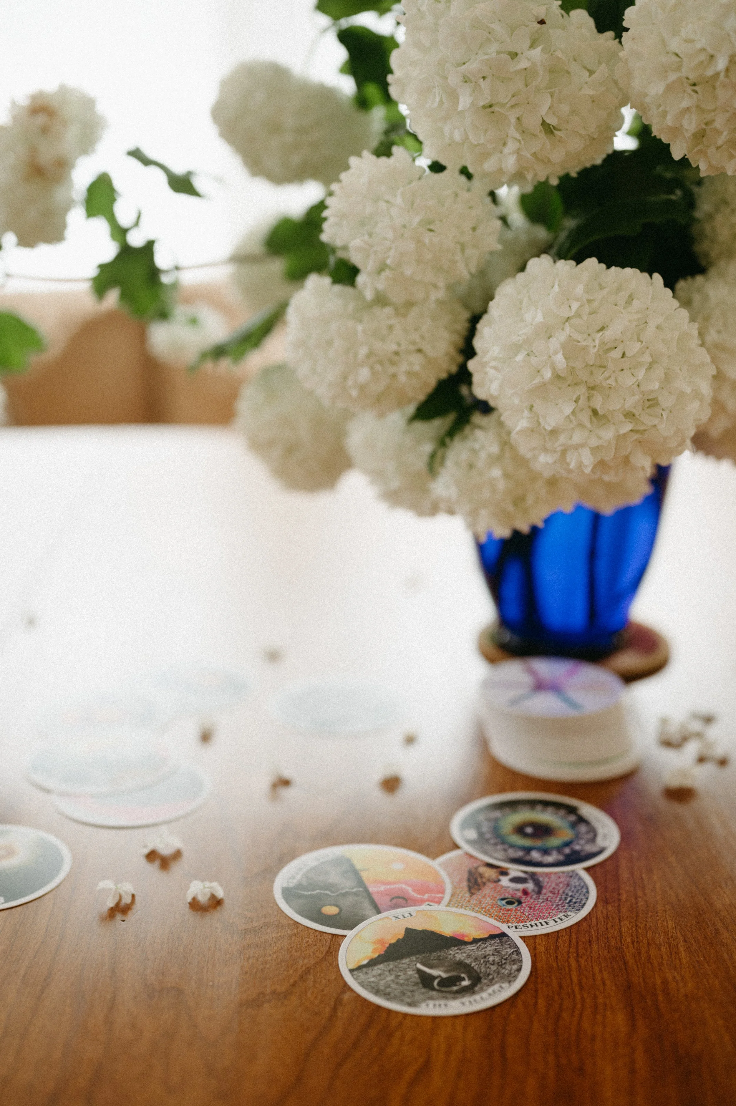 A blue glass vase with a bouquet of white hydrangea flowers. The vase is placed on a table with scattered decorative circular stickers featuring various landscape images and designs. Some flowers are visible in the background.