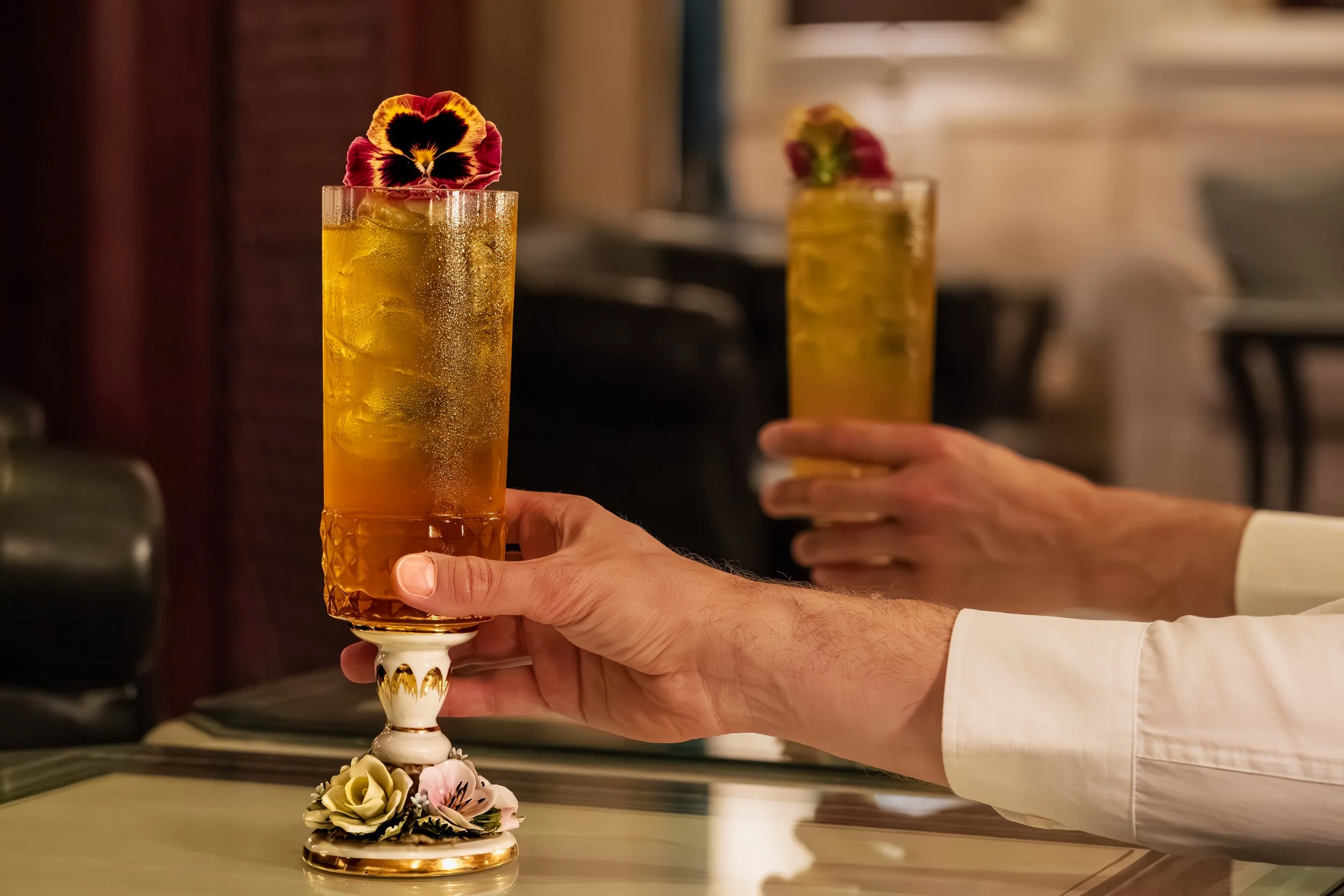 Il bicchiere contiene un cocktail arancione con ghiaccio, decorato con un fiore commestibile, tenuto da una mano su una base elegante.