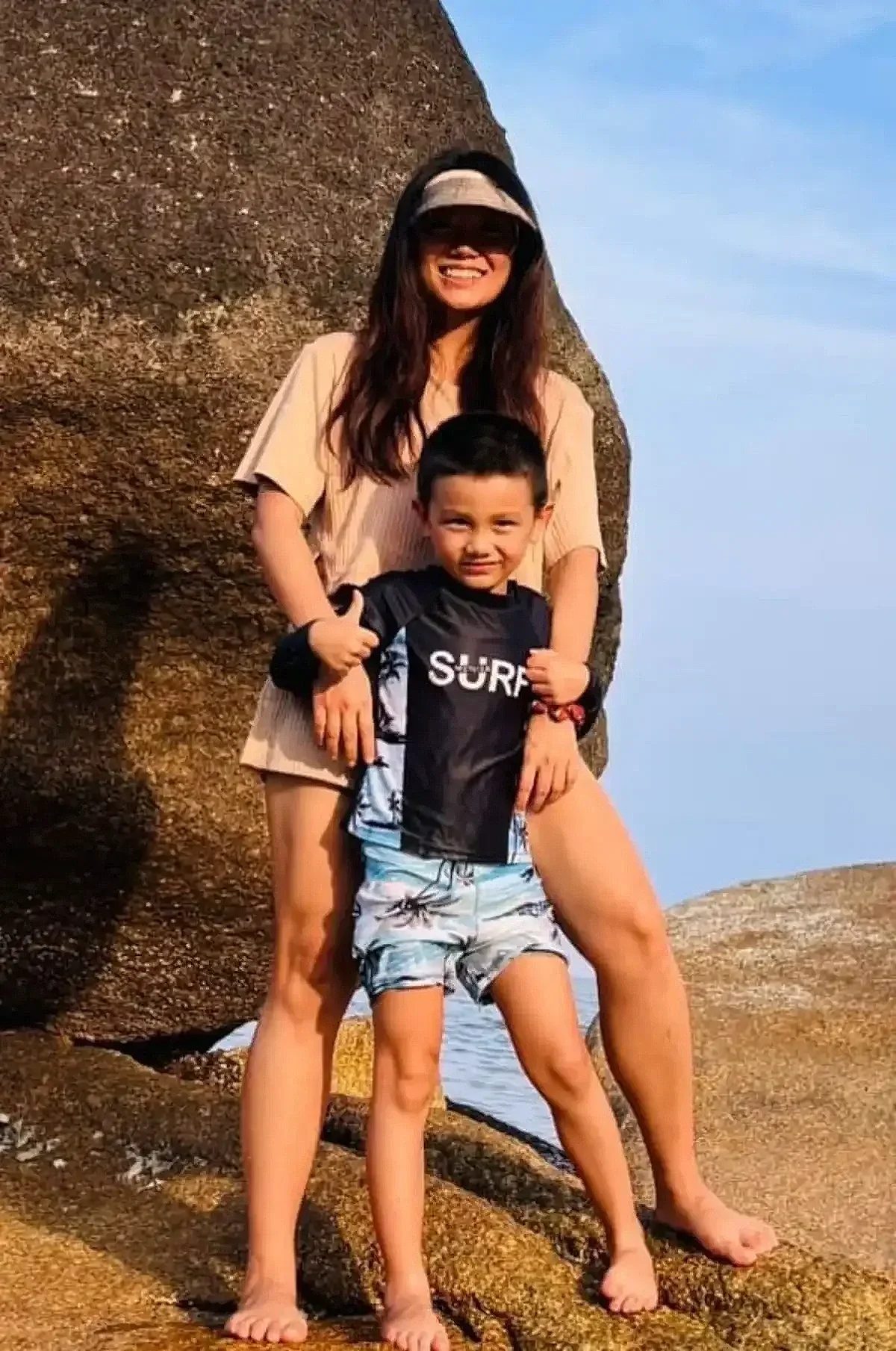 A woman and a young boy standing on rocks near the water at the beach, both smiling. The woman is wearing sunglasses and a beige outfit, while the boy is in a black surf shirt and light blue shorts.