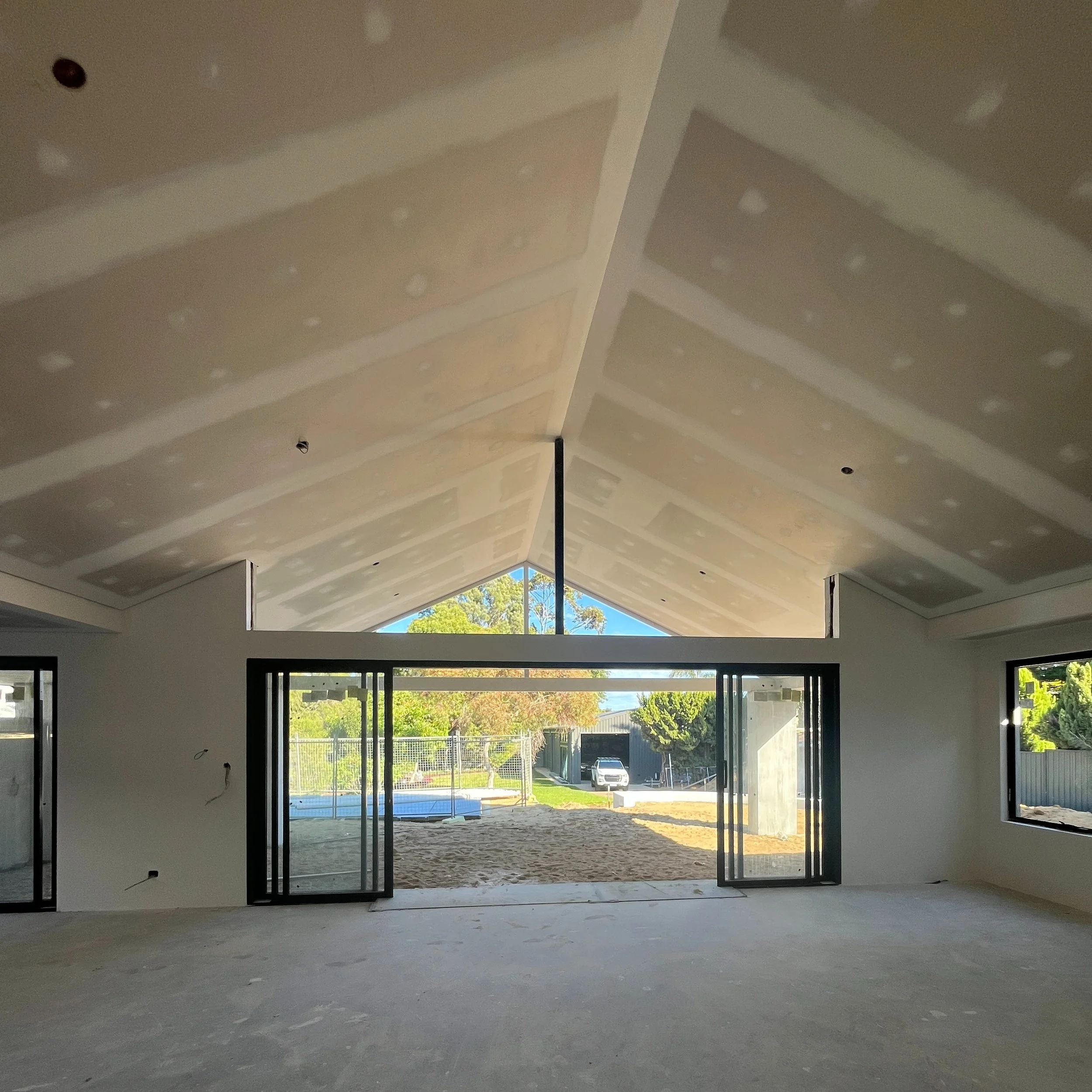 Interior of a house under construction with unfinished drywall on the ceiling and large sliding glass doors opening to a yard.