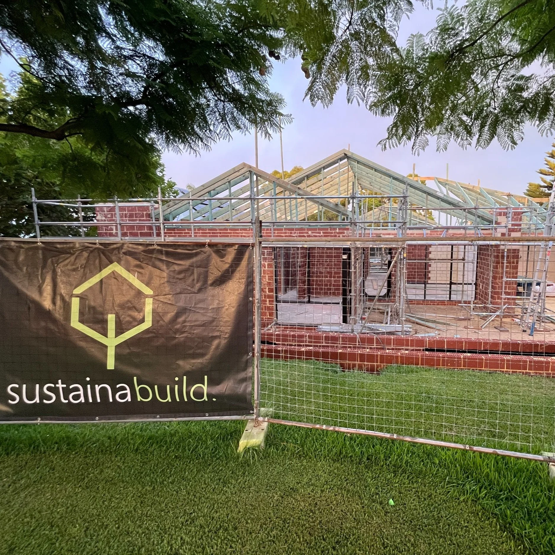A house under construction with scaffolding and a partially completed brick exterior, surrounded by green grass and trees, with a banner that reads 'sustainabuild.'