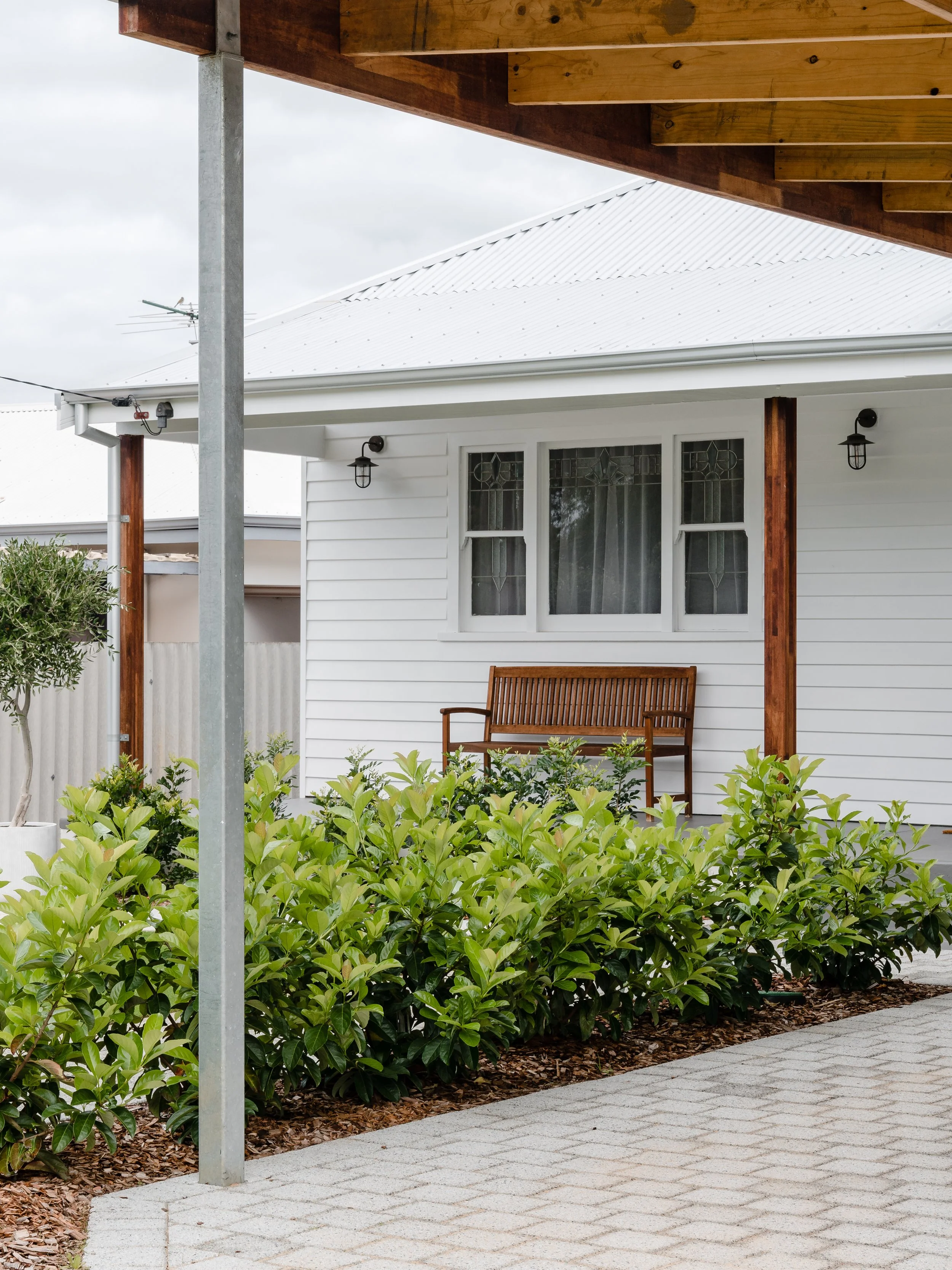 Front porch of a white house with a wooden bench, two windows with stained glass, black outdoor lights, and green bushes in the garden, with a tree on the left and a paved sidewalk.
