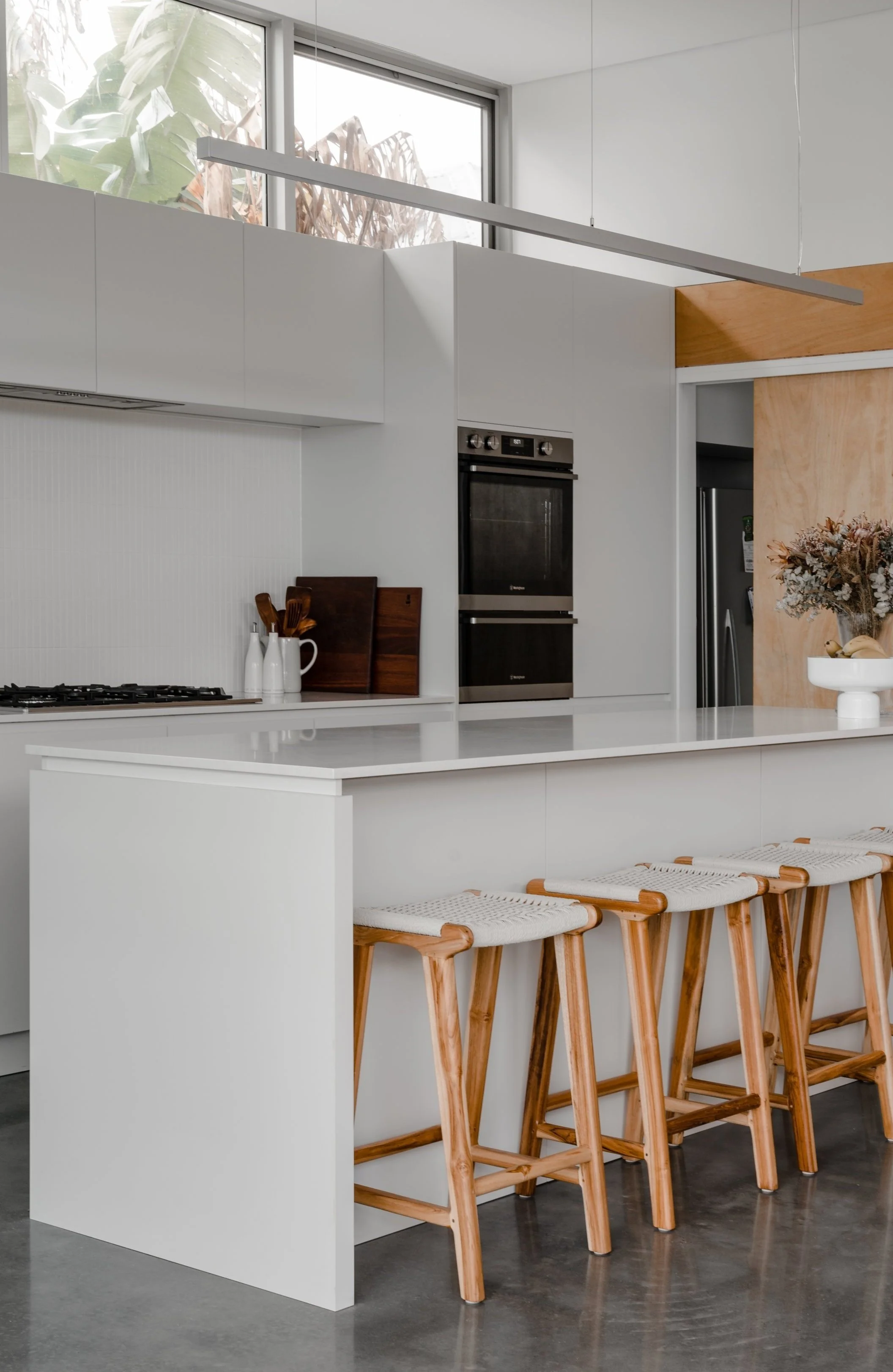 Modern white kitchen with a breakfast bar, wooden stools, built-in oven, and a large window with greenery outside.