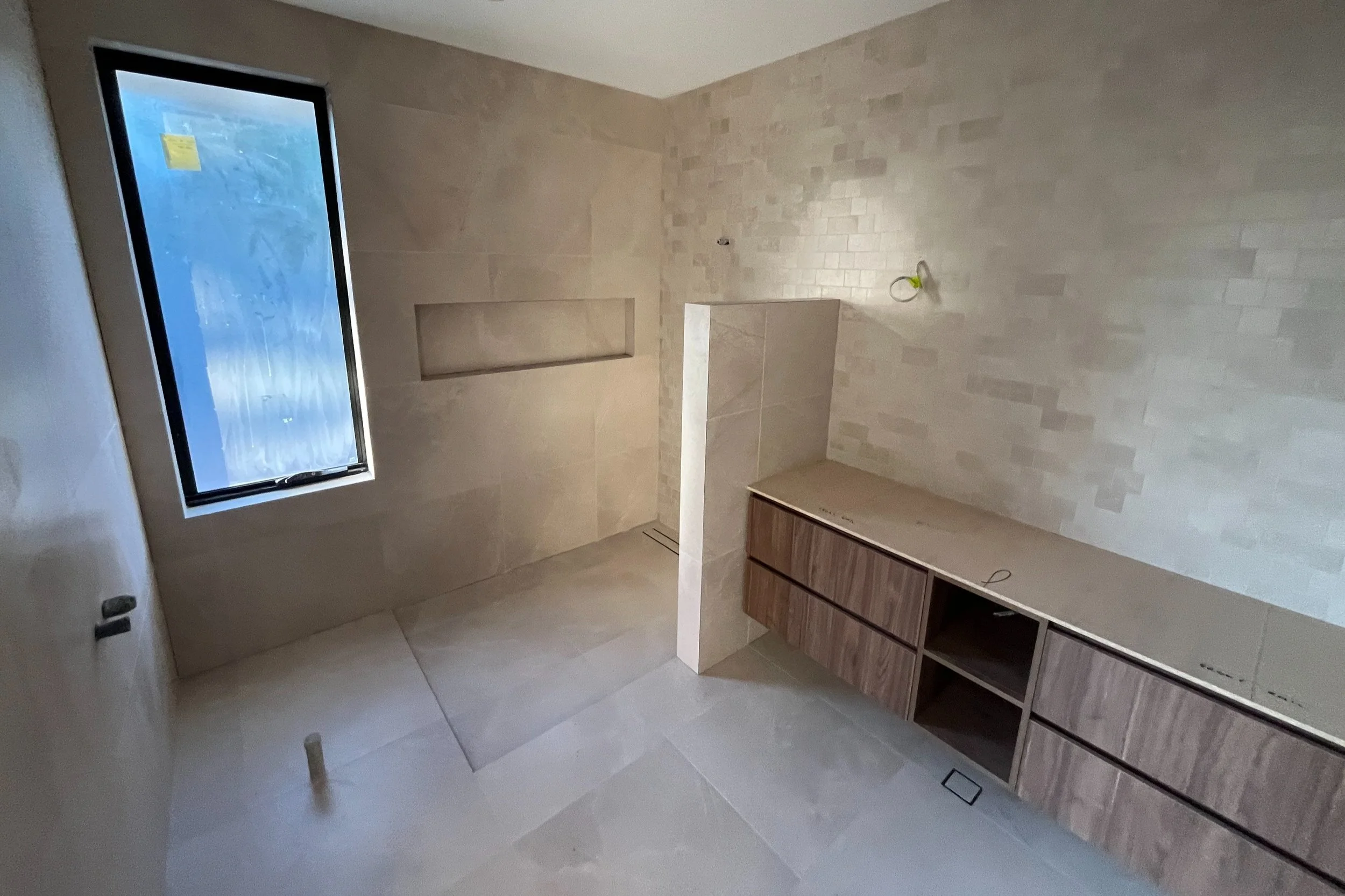 An unfinished bathroom with beige tiled walls and flooring, a window on the left, a built-in shower niche, and a wooden vanity or cabinet on the right with electrical wiring visible.