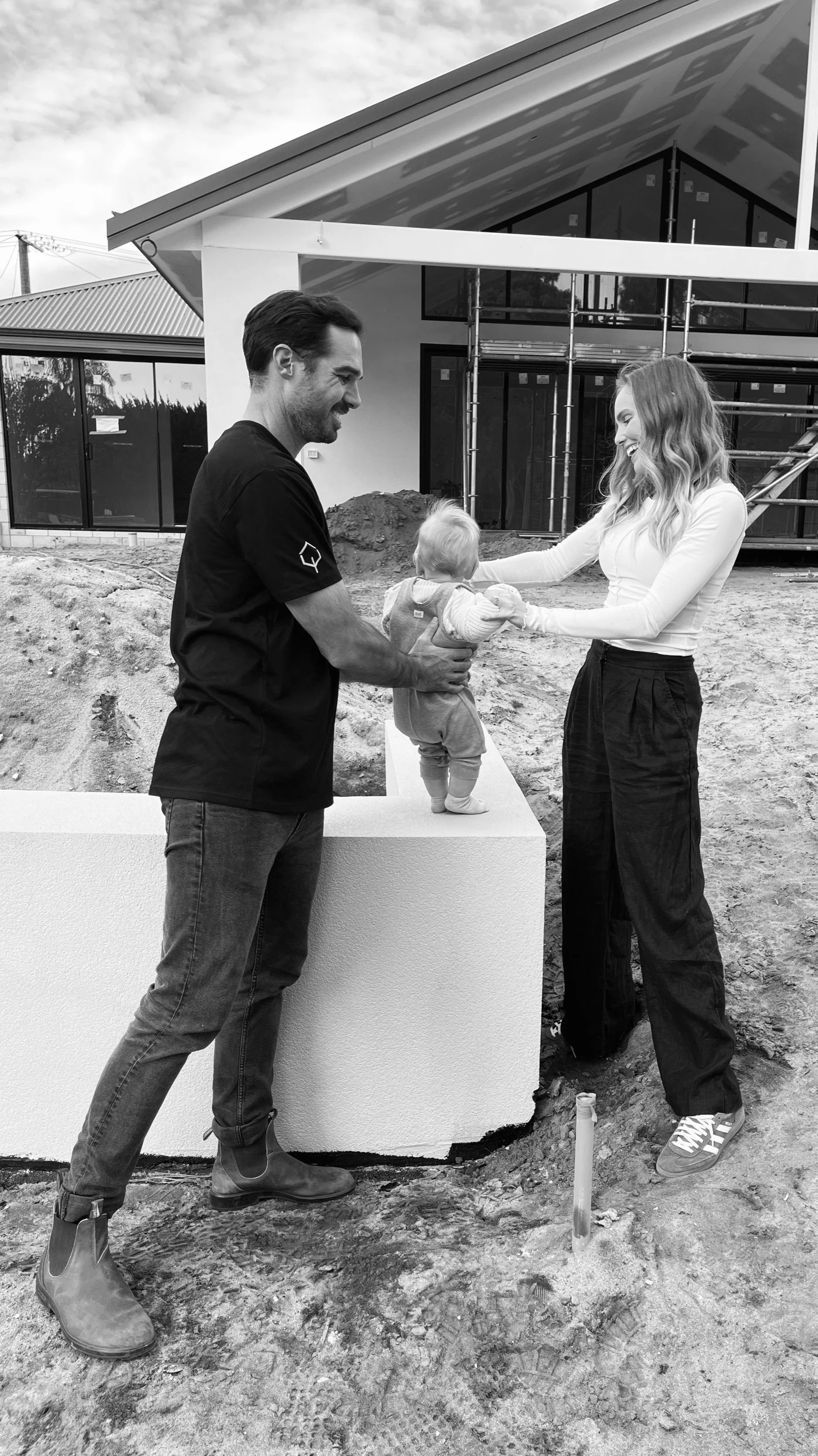 A couple with a small child on a construction site, smiling and holding the child's hands, in front of a new house under construction.