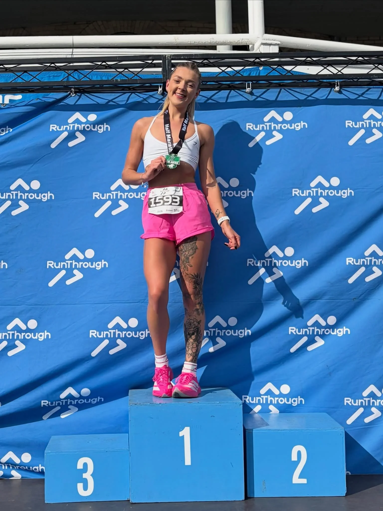 A woman standing on the first place podium at a race event, holding a medal, wearing athletic attire, pink shorts, and pink running shoes, with a background banner displaying the 'Run Through' logo.