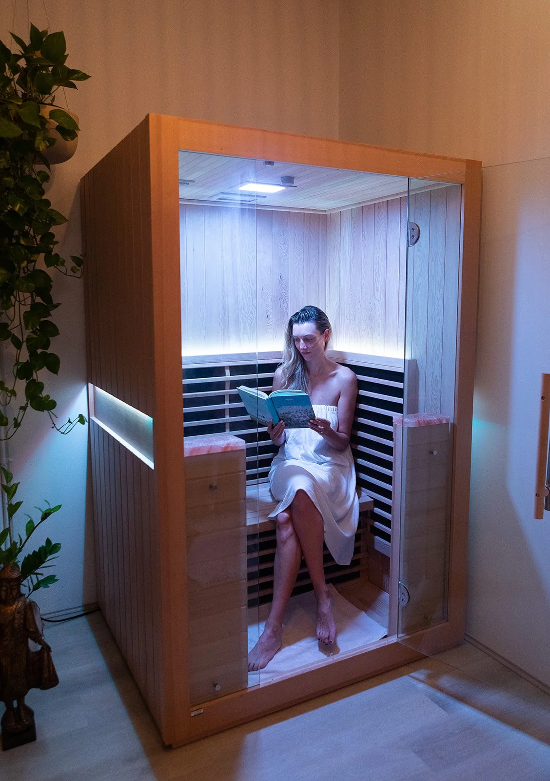 A woman in a towel sitting inside a wooden sauna reading a book.