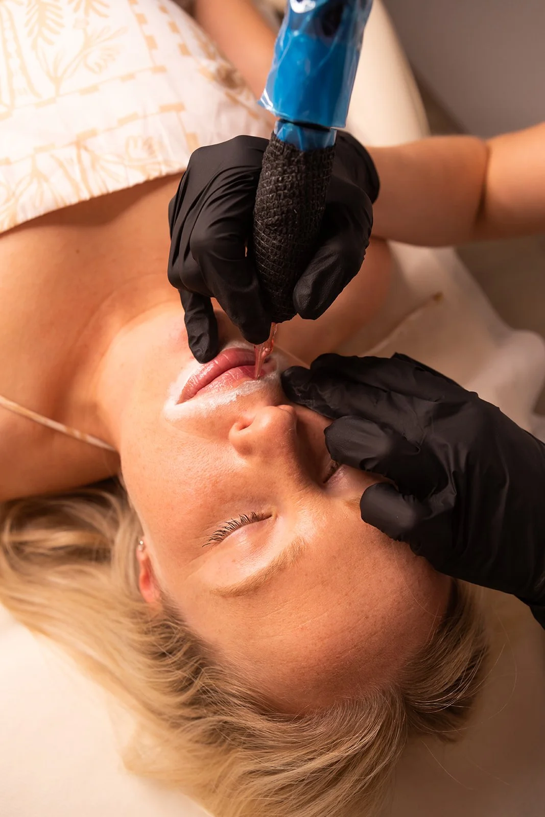 A woman lying down with her eyes closed as a tattoo artist using black gloves applies or touches up a lip tattoo with a tattoo machine.