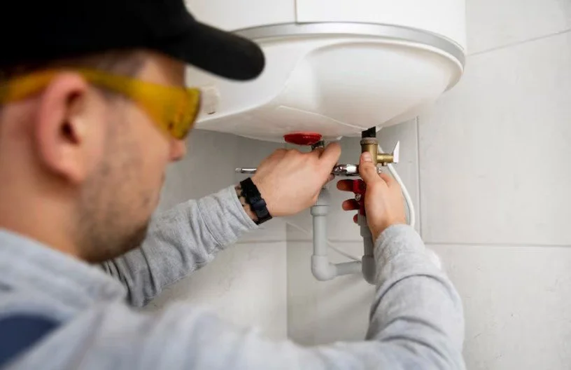 A person wearing safety goggles and a baseball cap working on plumbing underneath a bathroom sink.