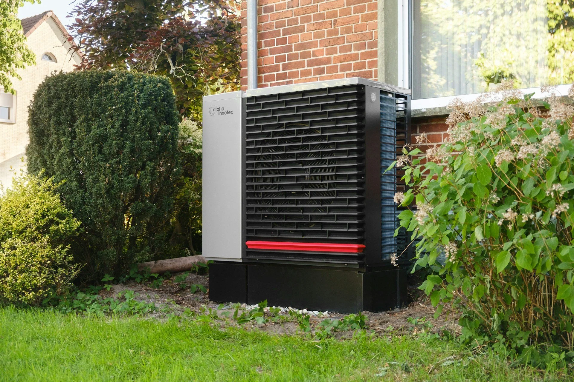 Outdoor air conditioning unit next to a brick house, surrounded by green plants and shrubs.