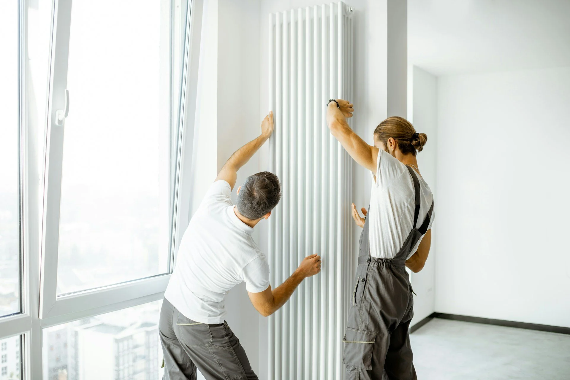 Two men and one woman install or inspect a vertical radiator near a window in a bright, modern room.