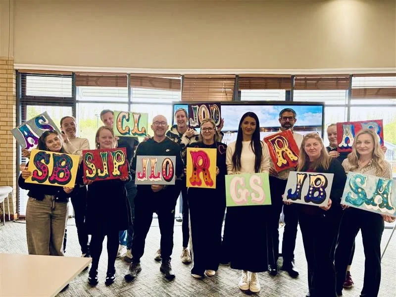 Group of people holding colorful decorative signs with letters and symbols in an indoor setting with large windows and a television screen in the background.