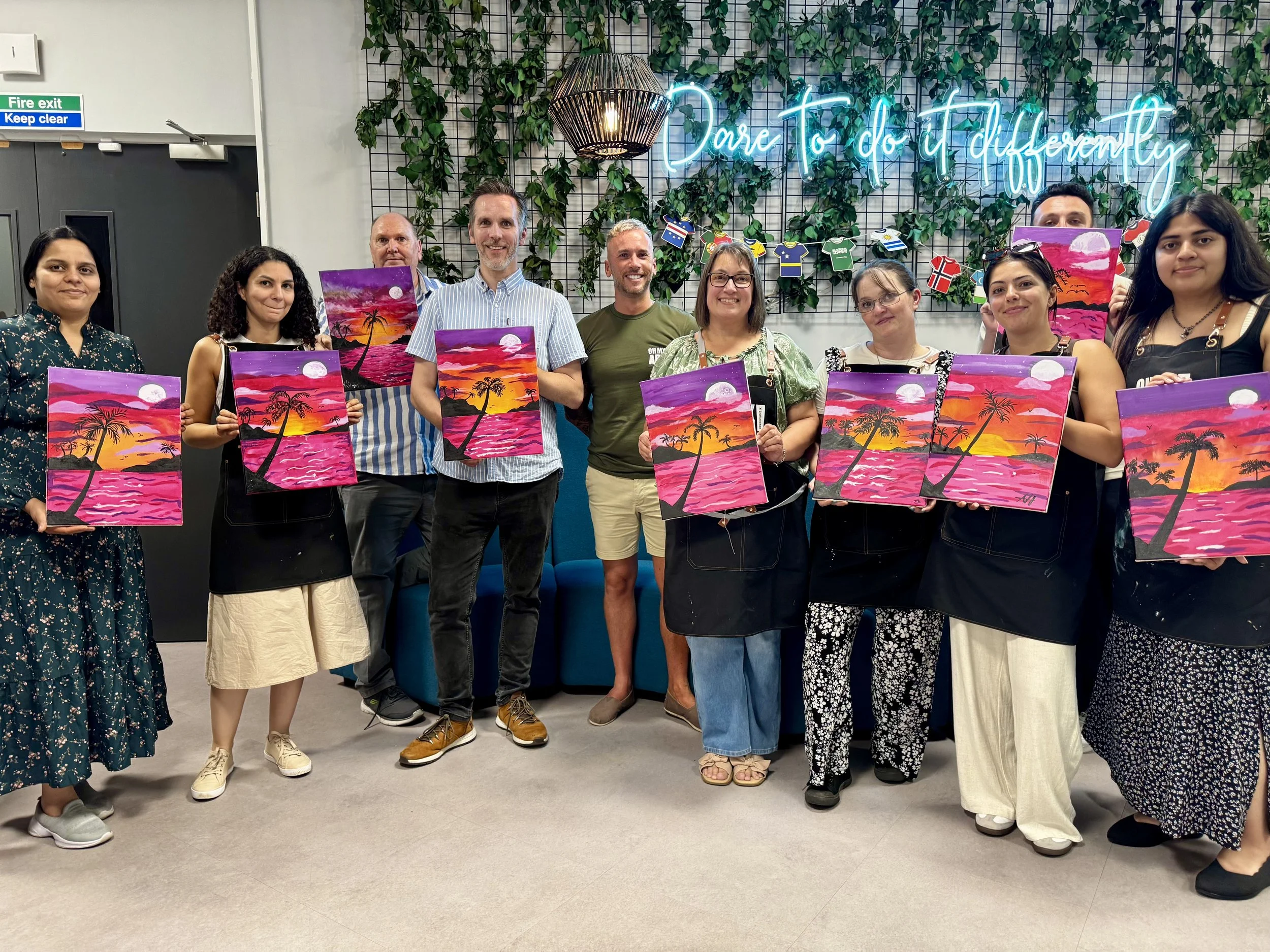 Group of people holding colorful sunset paintings with palm trees, standing in front of green leafy wall with neon sign that says 'Dare to do different'.