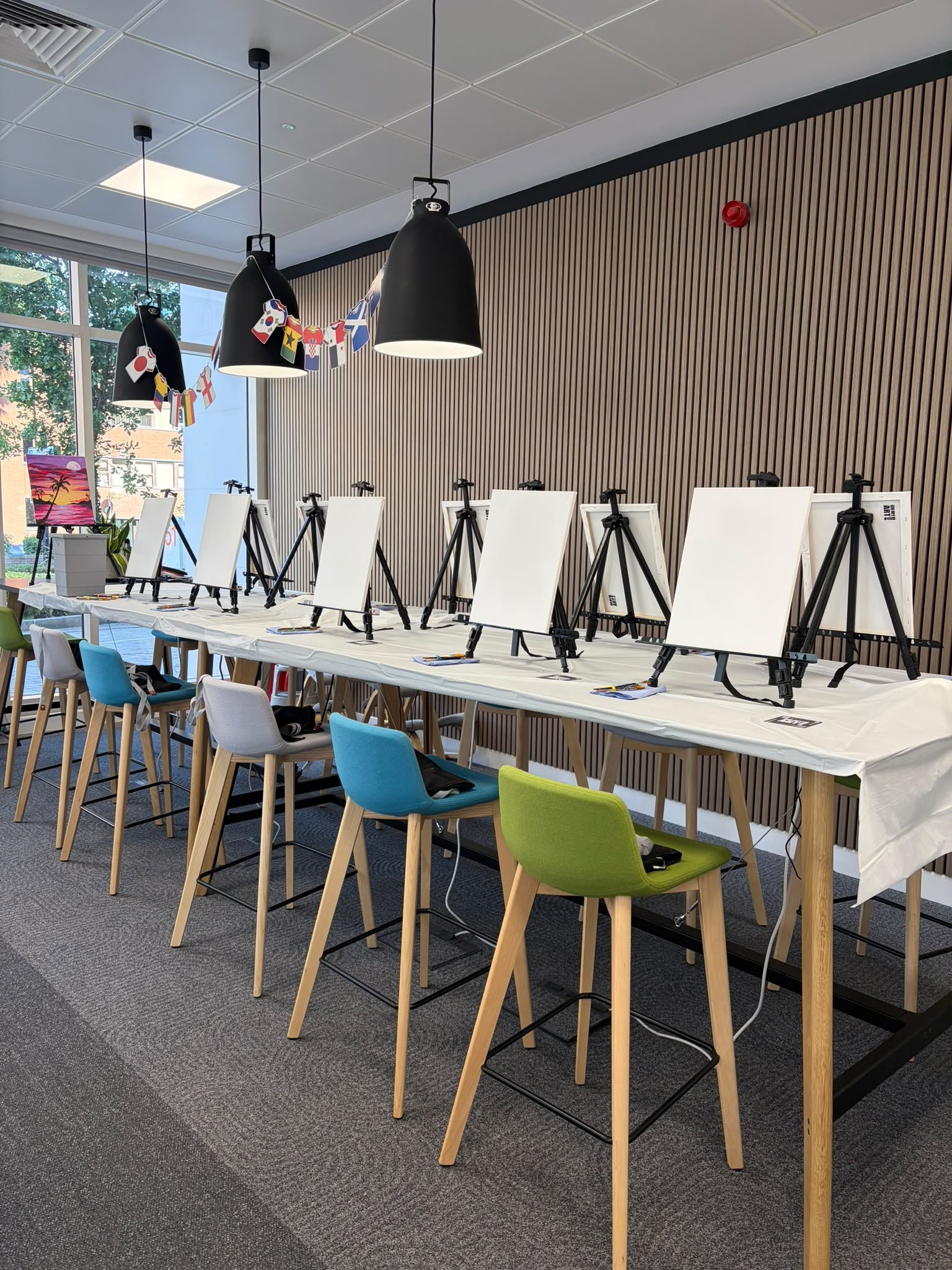 A room arranged for a painting or art event, with blank canvases on easels set up on a long table, chairs around it, and a window showing trees outside. There are hanging lights and a decorative banner with international flags.