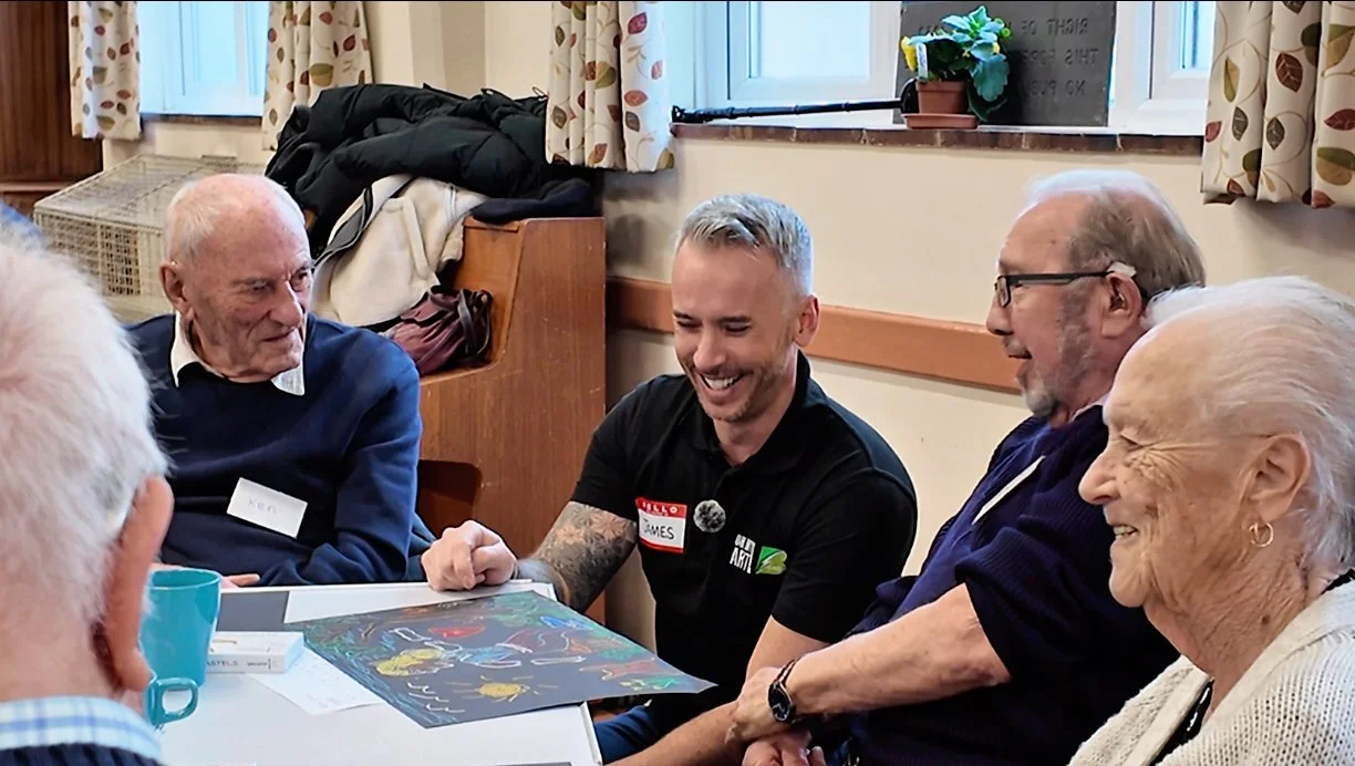 A group of five elderly and middle-aged people sitting at a table, smiling and interacting with each other. They appear to be engaged in a cheerful conversation or activity in a room with a window, floral curtains, and a small potted plant.
