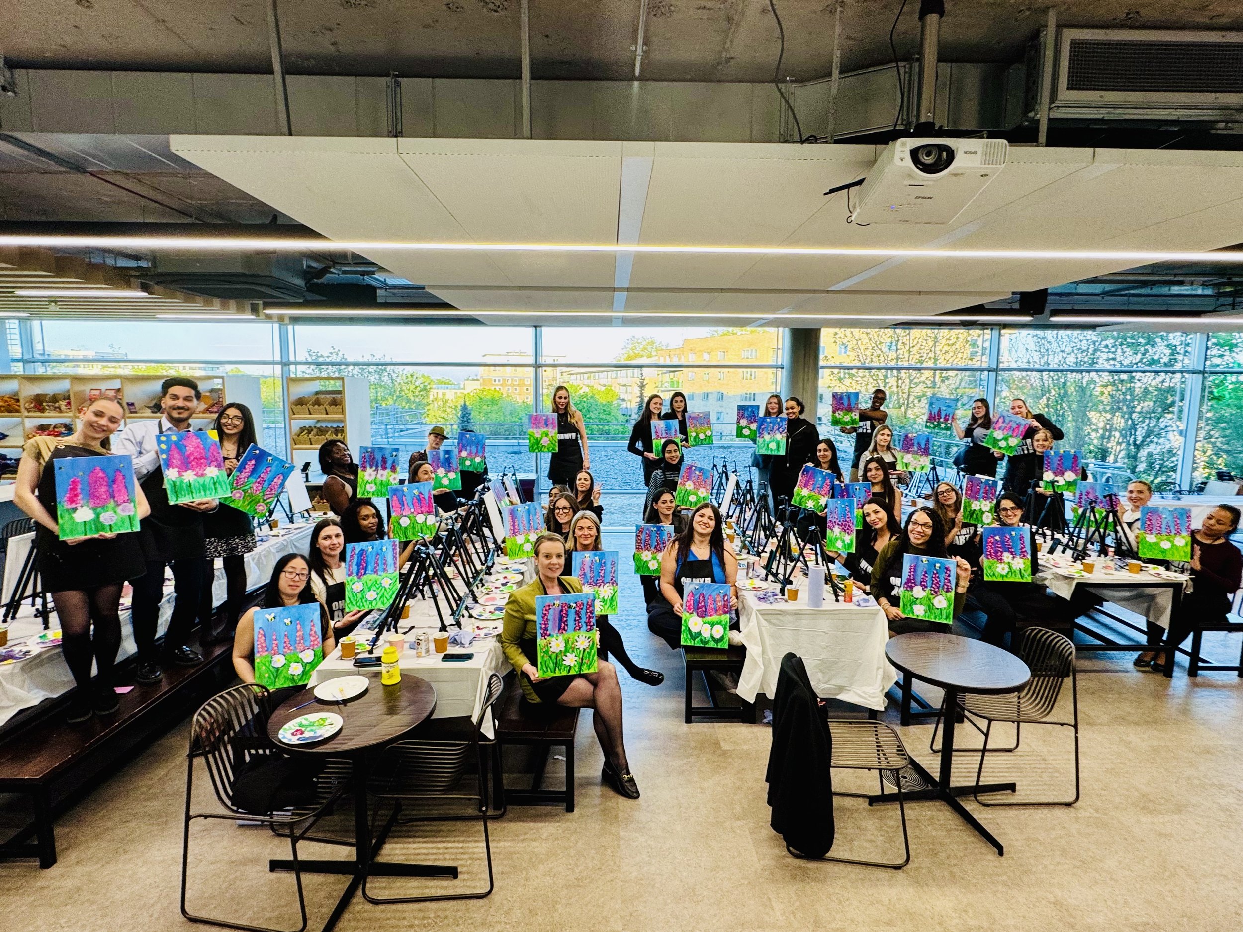 Group of people participating in a painting class, holding up their colorful artwork of pink and purple flowers on a blue background, in a bright, modern indoor space with large windows.