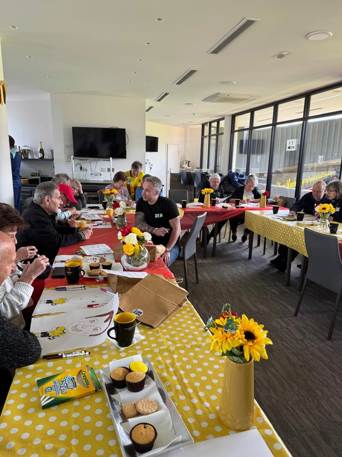 People gathered around tables with yellow polka dot tablecloths, decorated with sunflower centerpieces, participating in a community event or meeting in a bright, modern room with large windows, a TV, and a kitchenette in the background.
