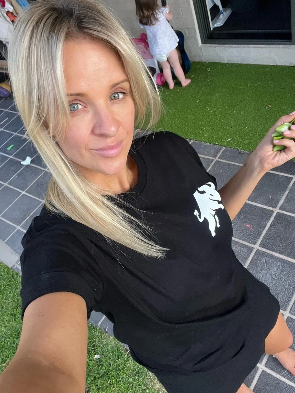 A woman with blonde hair, wearing a black shirt, taking a selfie outdoors on a patio area with artificial grass and tile flooring. There are children in the background.