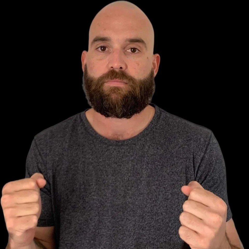 A man with a bald head and full beard looks directly at the camera with a serious expression, wearing a dark grey t-shirt and making fists with both hands against a black background.