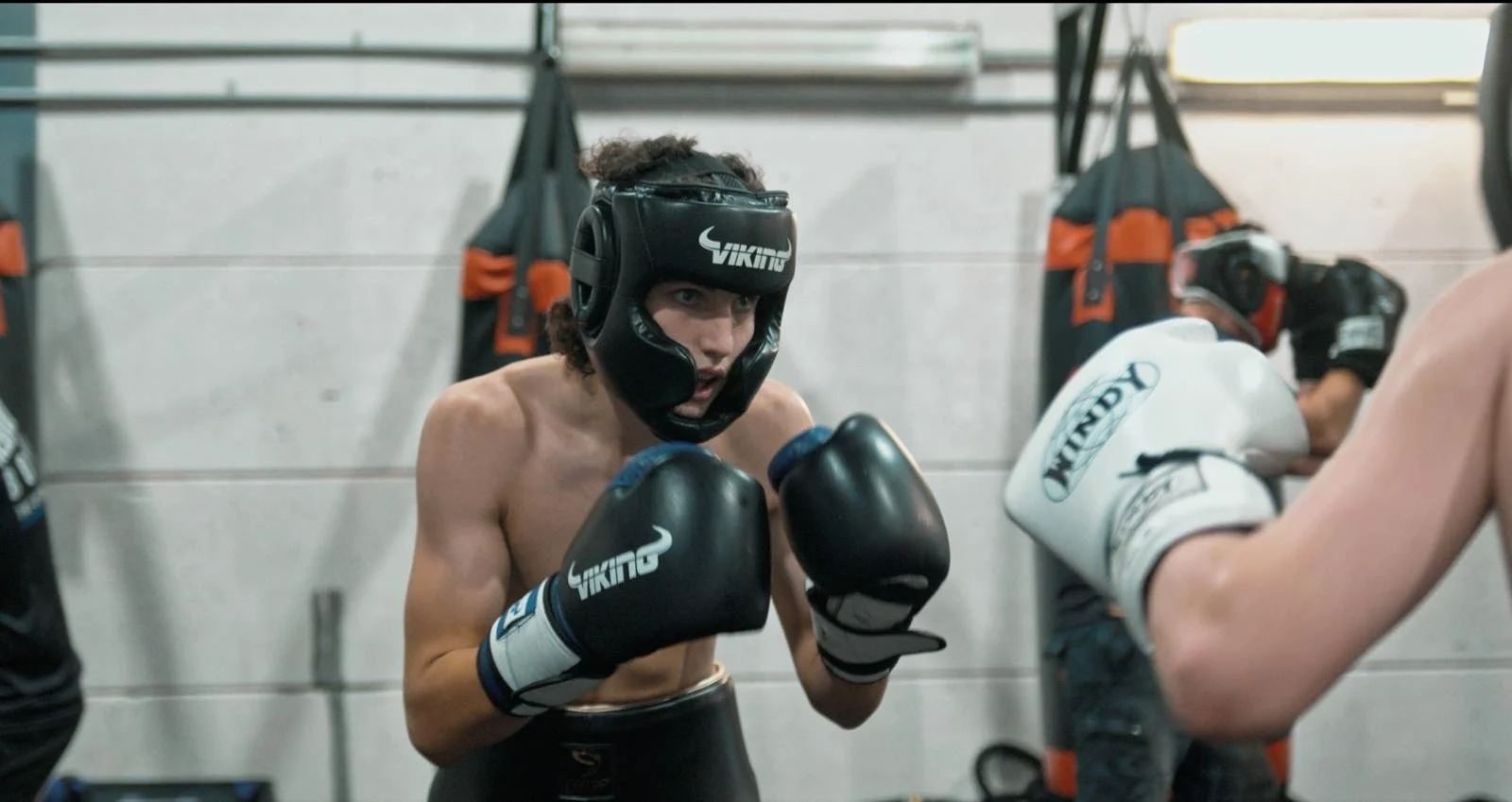 Junior Boxing – The Foundation of Boxcamp , A young shirtless male boxer with curly hair wearing a black helmet and black gloves, practicing boxing with a partner in a gym.