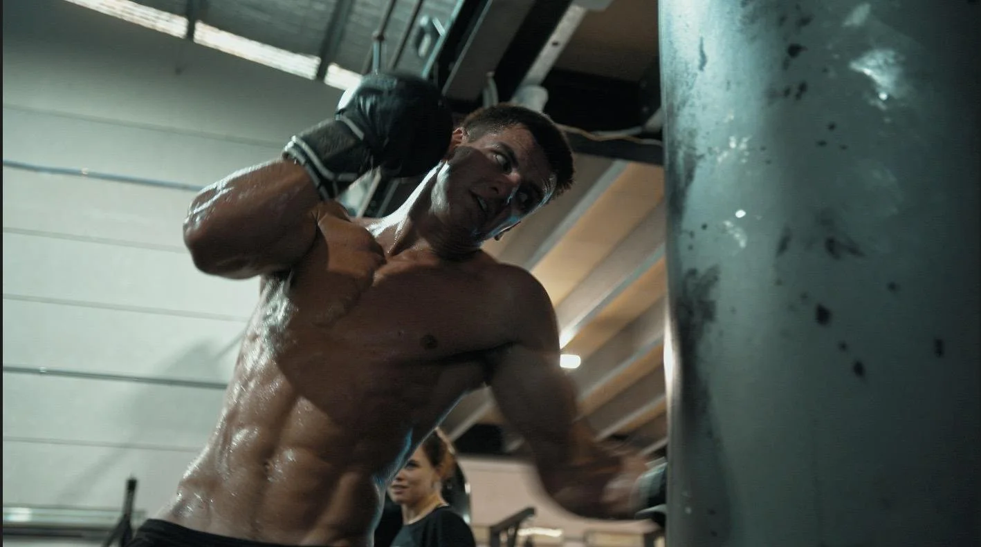A muscular man with wet skin, shirtless, wearing workout gloves, exercising at the gym by punching a heavy bag with a determined look.