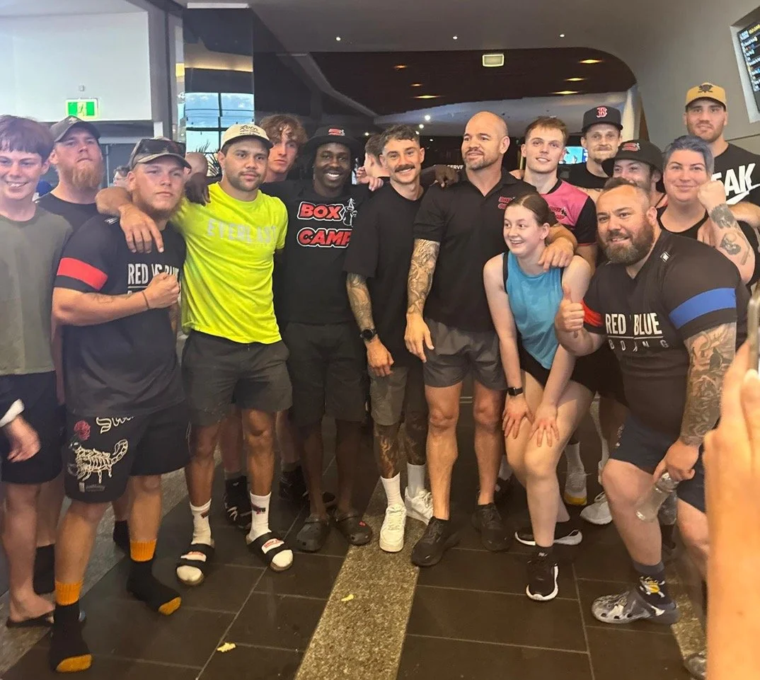 Group of people, mostly young men, posing together indoors, some wearing athletic attire, smiling and showing their fists, suggesting a sports or fitness event.