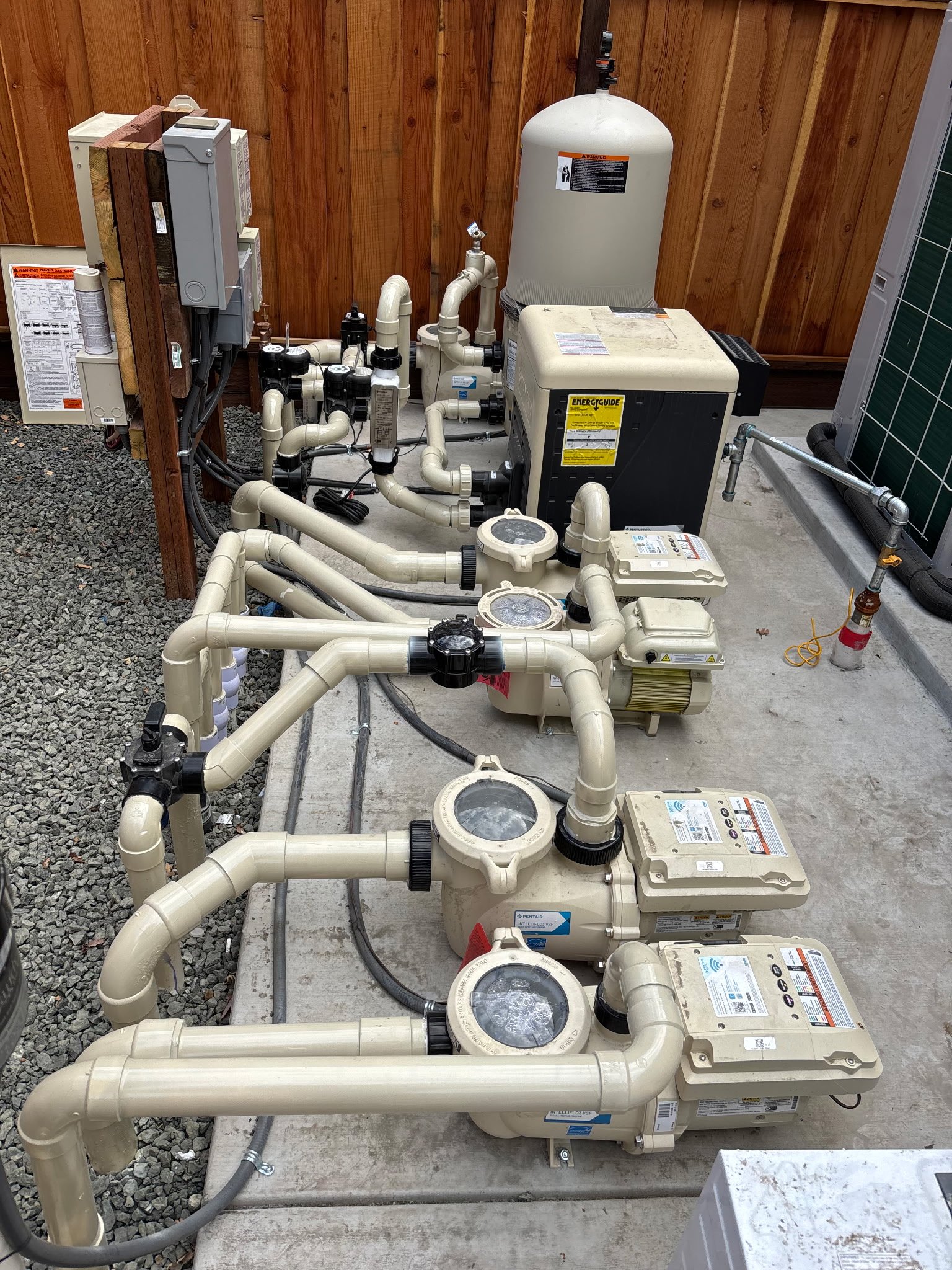 A pool pump and filtration system with pipes and valves installed on a concrete surface next to a wooden fence.