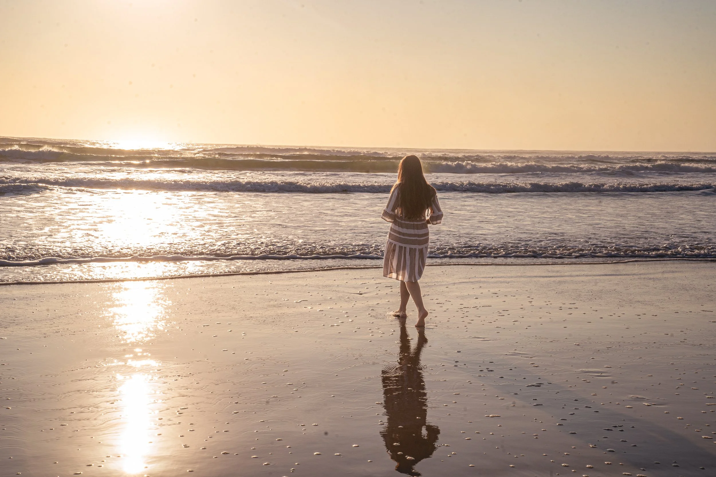 A woman with long hair standing on the beach facing the ocean during sunset, wearing a striped dress.
