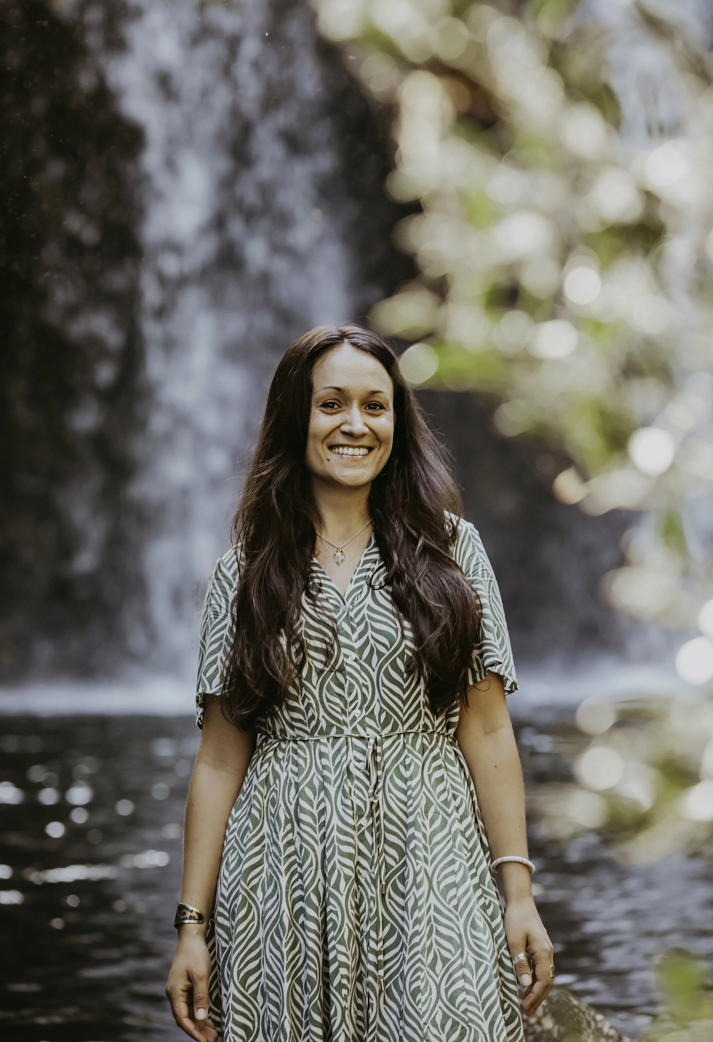 A woman with long brown hair and a patterned green and white dress standing near a waterfall with blurred bright foliage in the background.