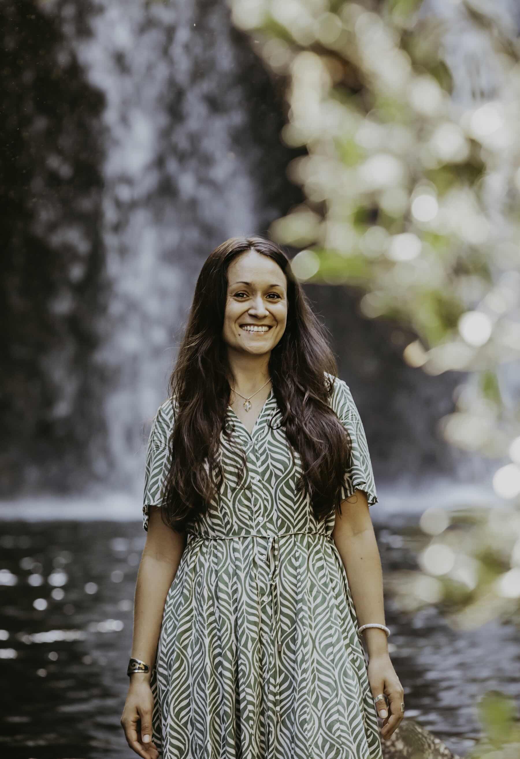 A woman with long dark hair wearing a patterned dress, standing in front of a waterfall with light bokeh.