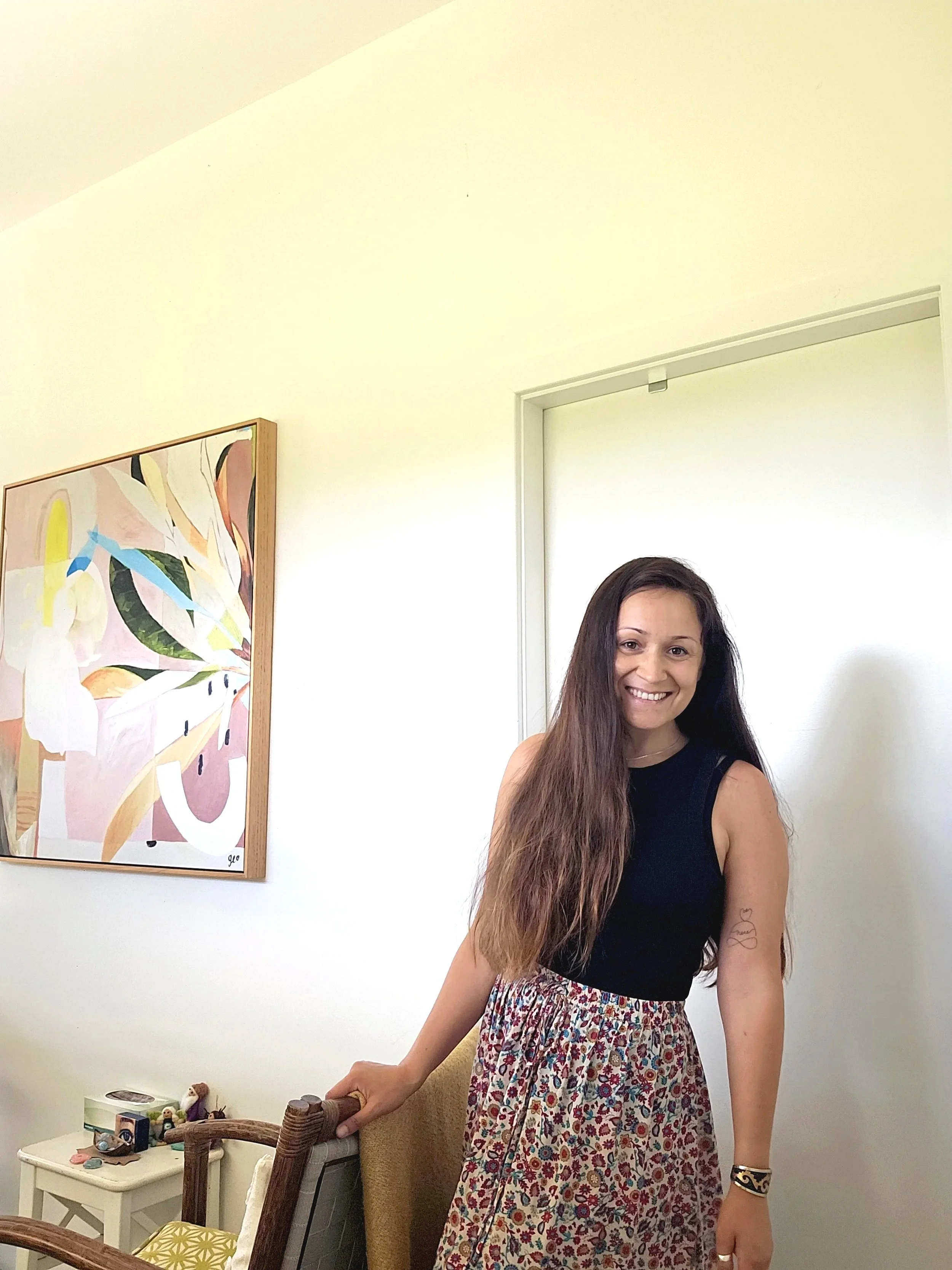 A smiling woman with long brown hair, wearing a black top, a floral skirt, and a bracelet, standing in a room with white walls and artwork, near a small table with decorative items.