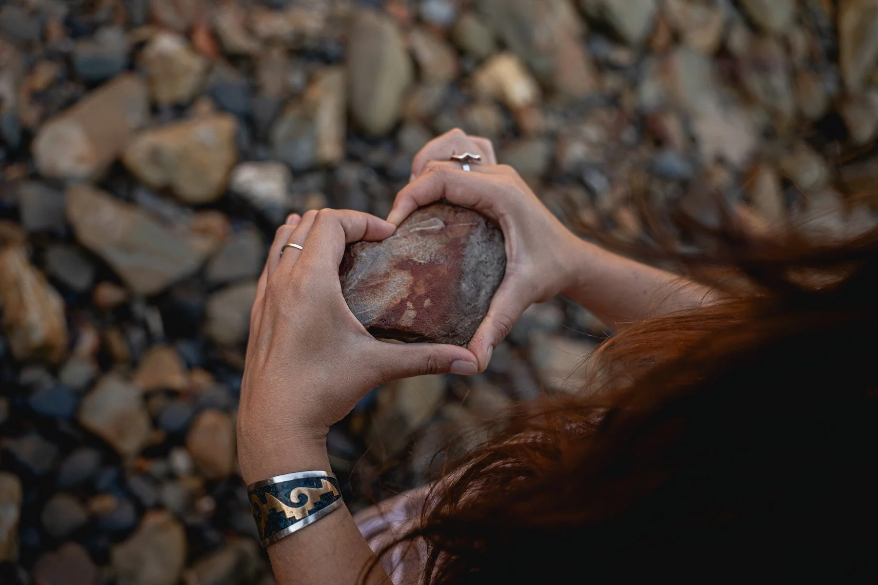 A person with red hair holding a reddish-brown rock with both hands, standing on a bed of smaller rocks.