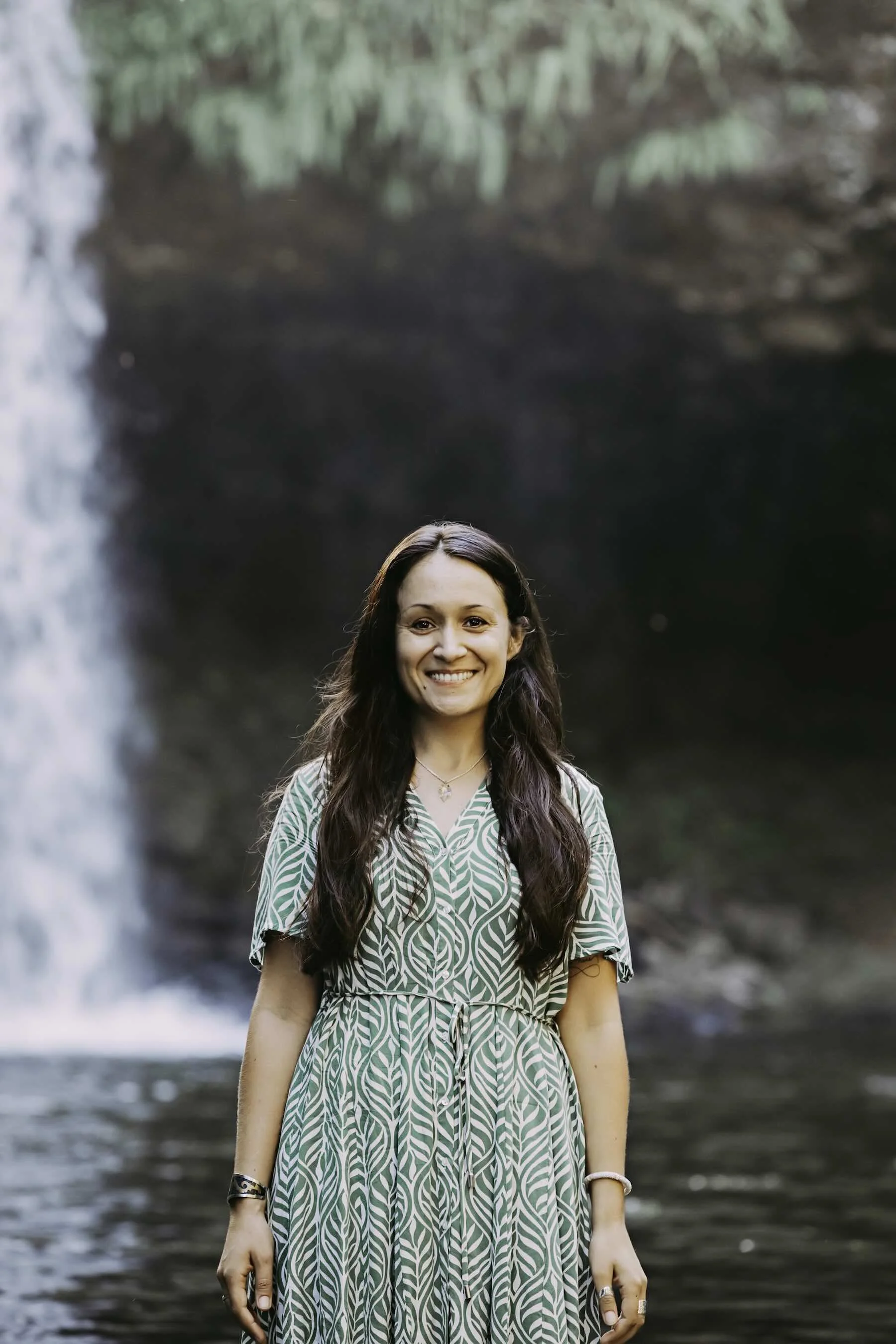 A woman with long brown hair, wearing a green patterned dress, standing in front of a waterfall with greenery in the background.
